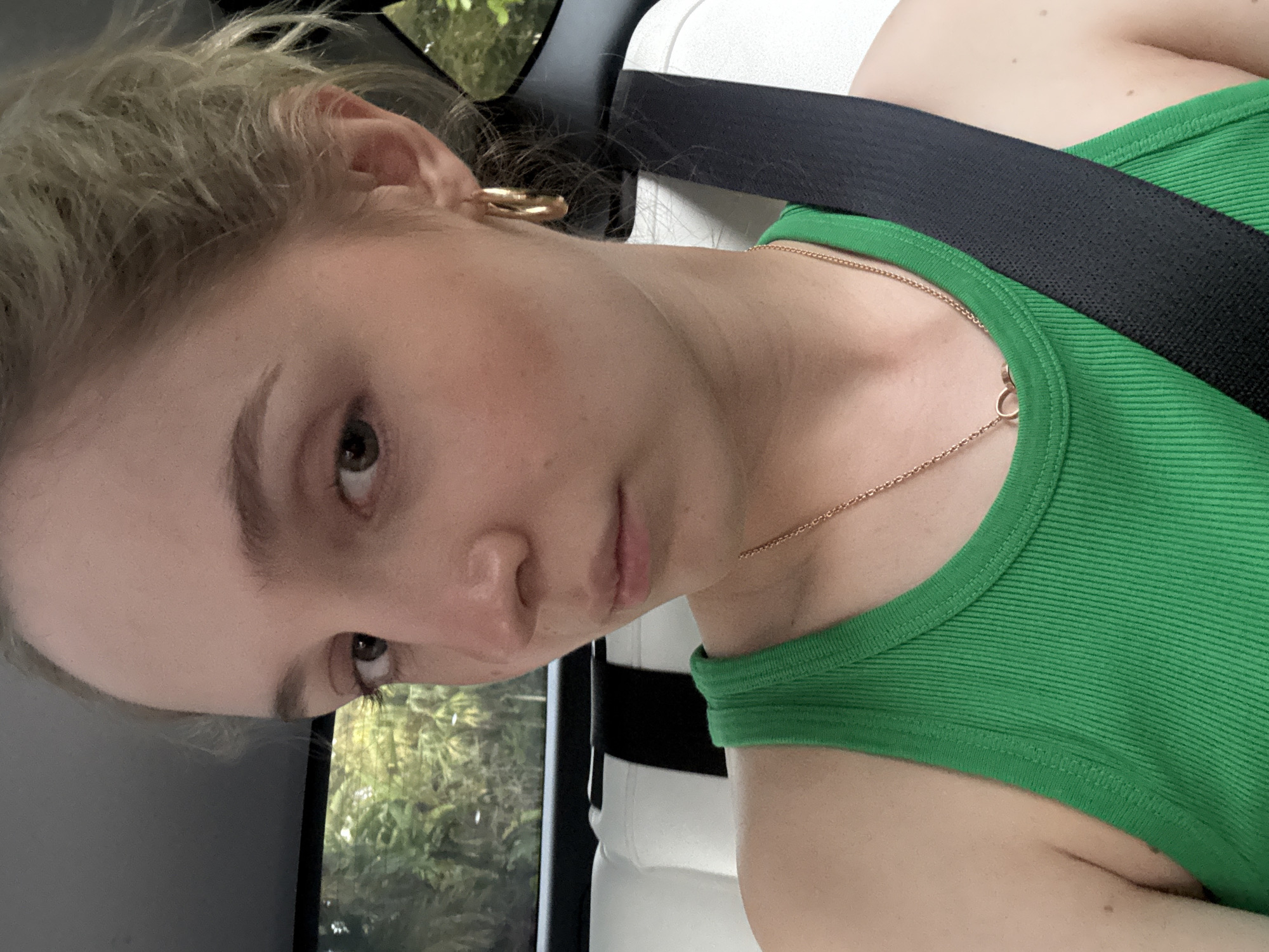 A young woman with blonde hair pulled back is seated in the passenger seat of a car, looking directly at the camera. She is wearing a green ribbed tank top and a gold necklace. A black seatbelt is visible across her chest. The interior of the car features white leather seats and a dark headliner. Outside the window, lush green foliage can be seen, suggesting a tropical environment. The lighting indicates it is daytime. The image does not provide specific details to determine the exact location within Miami, United States of America.