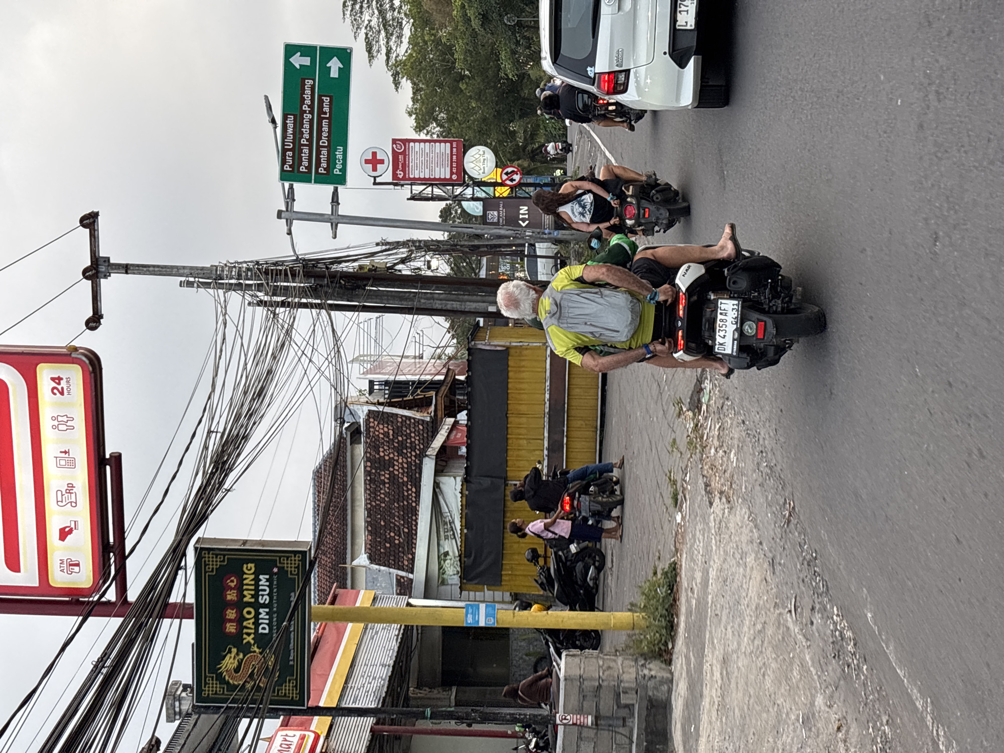 The image depicts a street scene in Kuta Selatan, Indonesia, characterized by a heavy density of overhead electrical wires. In the foreground, several individuals are riding motorcycles. The person closest to the camera, an older Caucasian male with white hair, is riding a white scooter, wearing a yellow and green shirt and a grey backpack. Behind him, a woman with dark hair, wearing a black tank top with a palm tree design and shorts, is riding on the back of a scooter. Further down the road, other motorcycles and a white car are visible.
To the left of the frame, a sign for "XIAO MING DIM SUM" with a dragon graphic is displayed prominently. Nearby, a red and white sign with a cross and text indicating "UNICARE" and various medical services is also visible. Green directional signs indicate "Pura Uluwatu", "Pantai Padang-Padang", "Pantai Dream Land", and "Pecatu". The environment suggests a bustling street with shops and businesses lining the road, indicated by awnings and building structures. The ground is paved with asphalt and concrete. The sky is overcast.