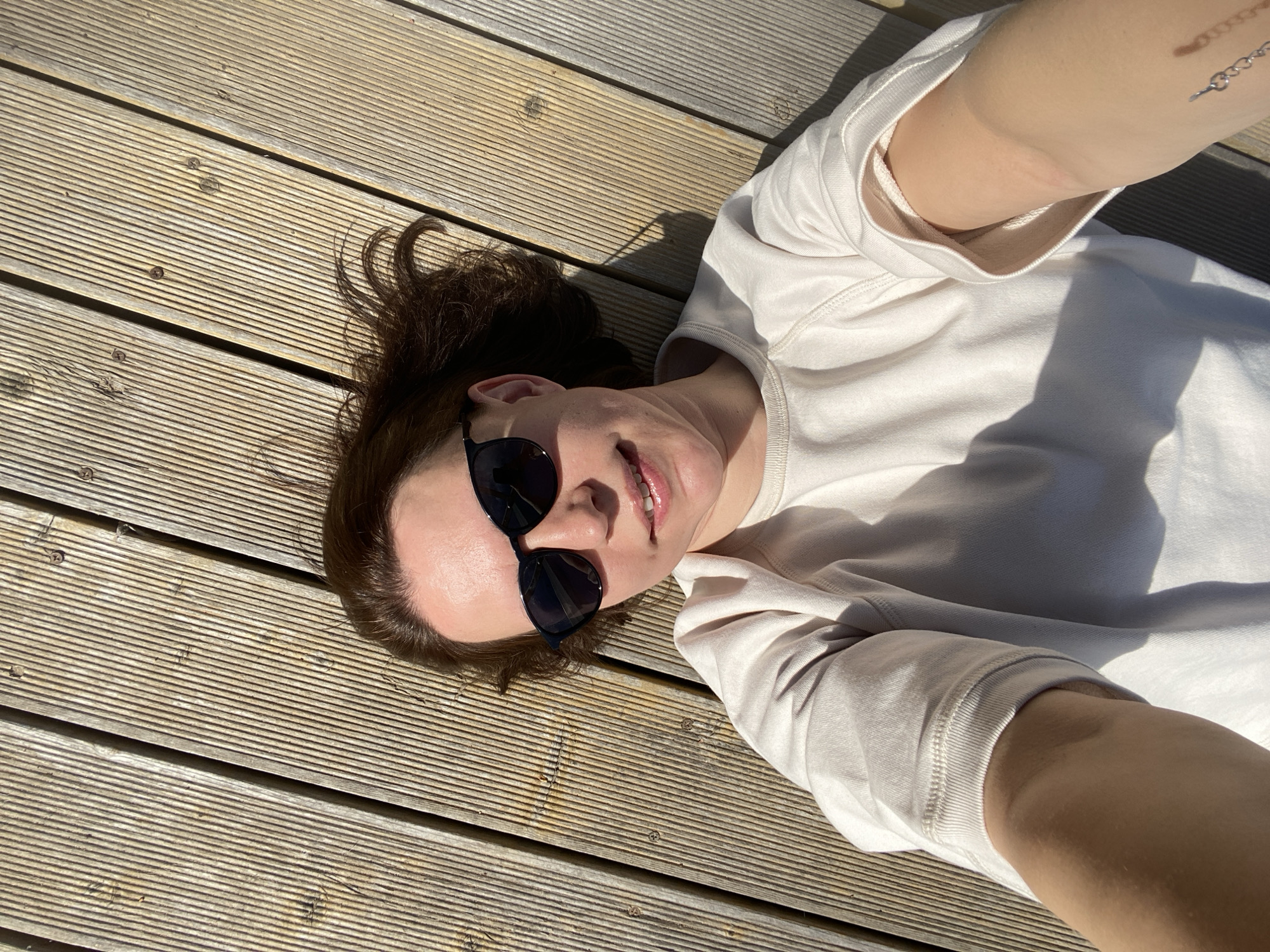 A woman lies on a wooden deck in the city of Chaikovsky, Russia. She wears a light-colored sweatshirt and sunglasses. Her head rests on the deck, and her arms are positioned around her head, suggesting she is taking a selfie. The wooden deck is made of evenly spaced planks, and the scene is bathed in sunlight. A small tattoo is visible on her right arm.