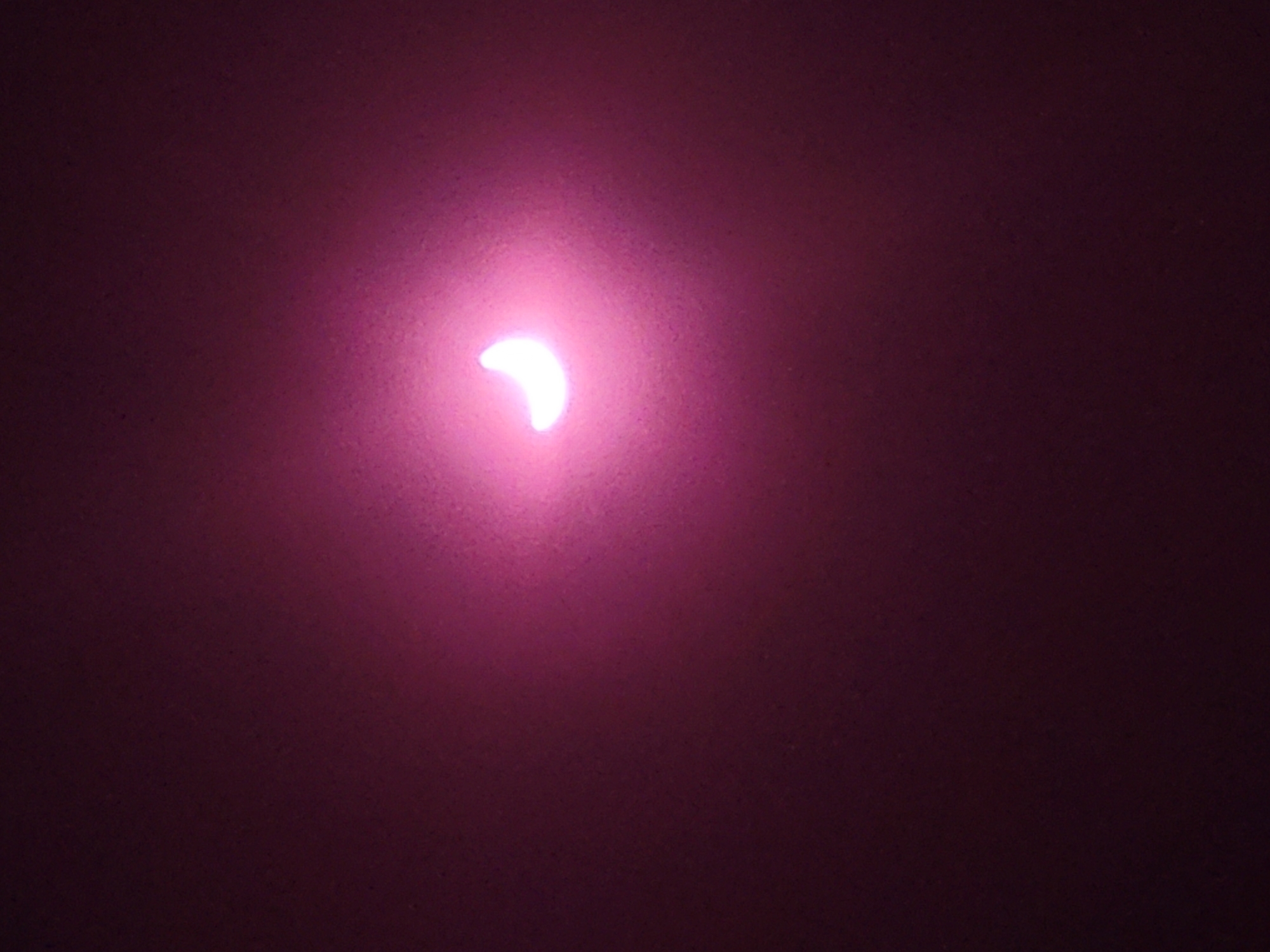 A crescent-shaped light, glowing with a pinkish hue, is visible against a dark, textured background. The light source appears to be a single, bright point, creating a soft glow around the crescent shape. The background is uniformly dark with subtle speckling or texture, possibly indicating a wall or fabric. The lighting suggests a low-light environment where the crescent is the primary illuminated object. There is no indication of a city, people, or any specific location.