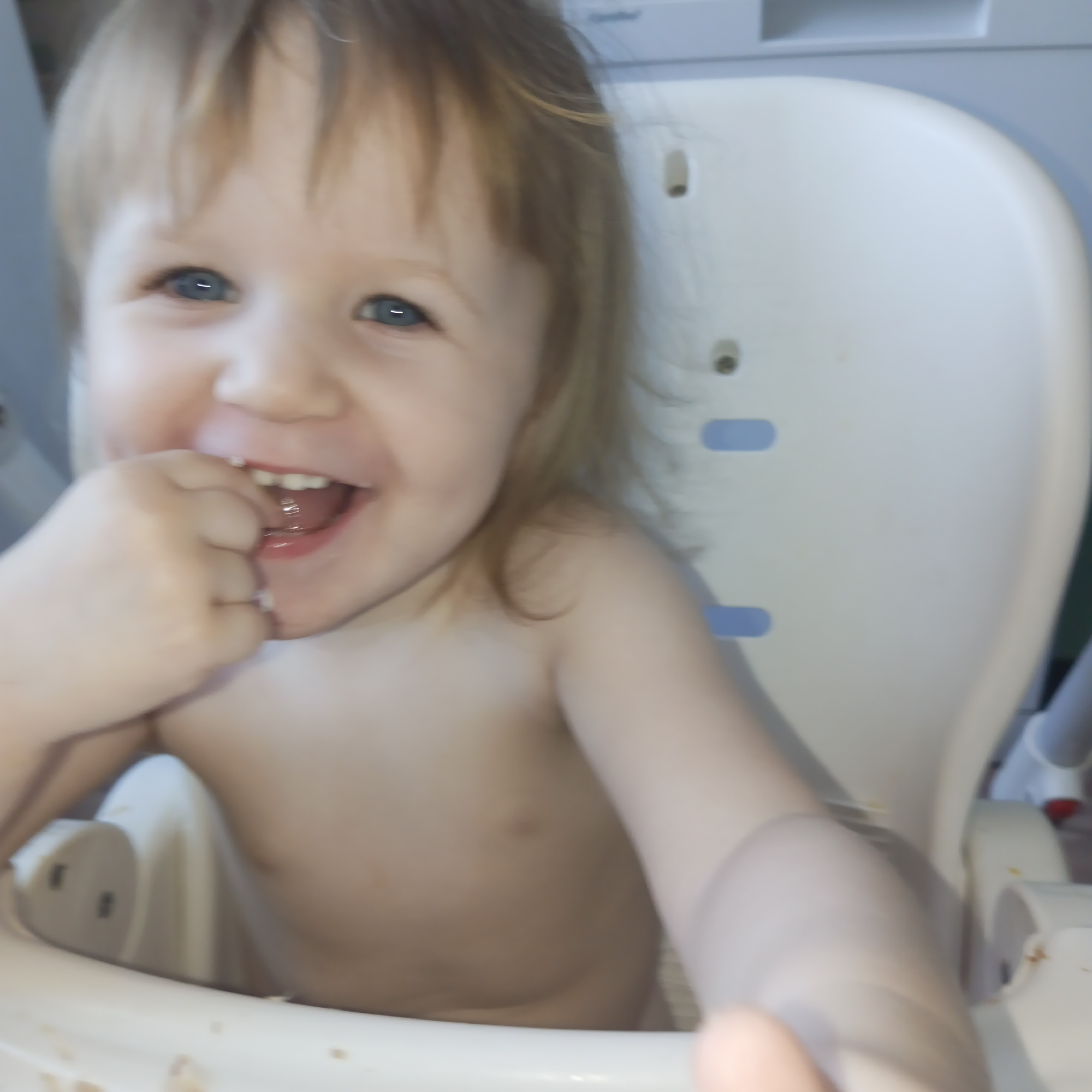 A toddler with light blonde hair and blue eyes is sitting in a white high chair. The child is smiling broadly with their mouth open, and they appear to be putting their fingers in their mouth. The child is shirtless. The high chair has blue accents. The background is blurry. No location information is available.