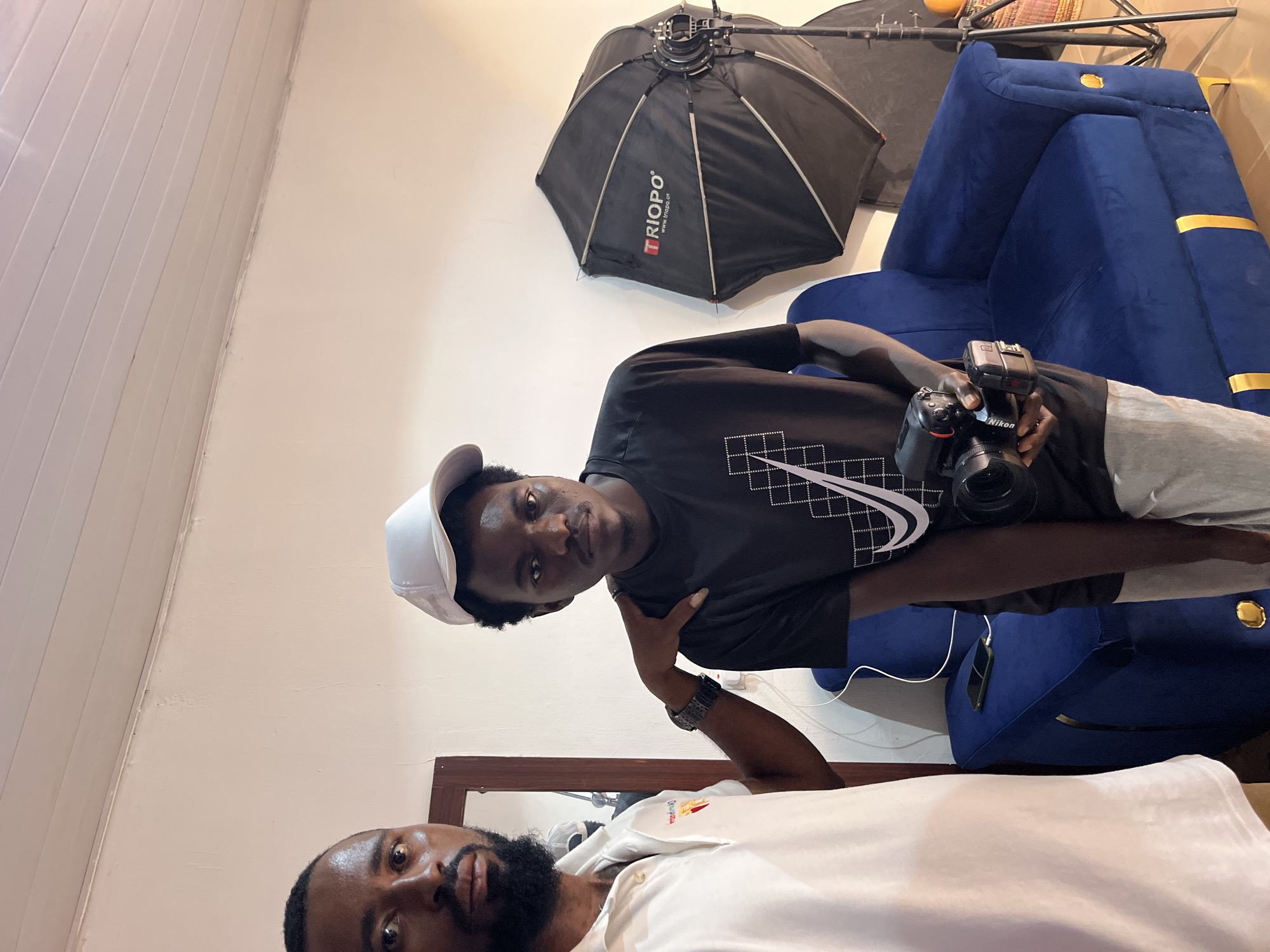 Two men are standing in a room. The man on the left is wearing a white polo shirt and has a beard. He has his hand on the shoulder of the man on the right. The man on the right is wearing a black t-shirt with a white Nike logo and a white baseball cap. He is holding a camera. A blue sofa is behind them, and a photography umbrella is to the right. A white cable is plugged into a phone on the sofa. The walls are white, and the ceiling is paneled. The floor is not visible.