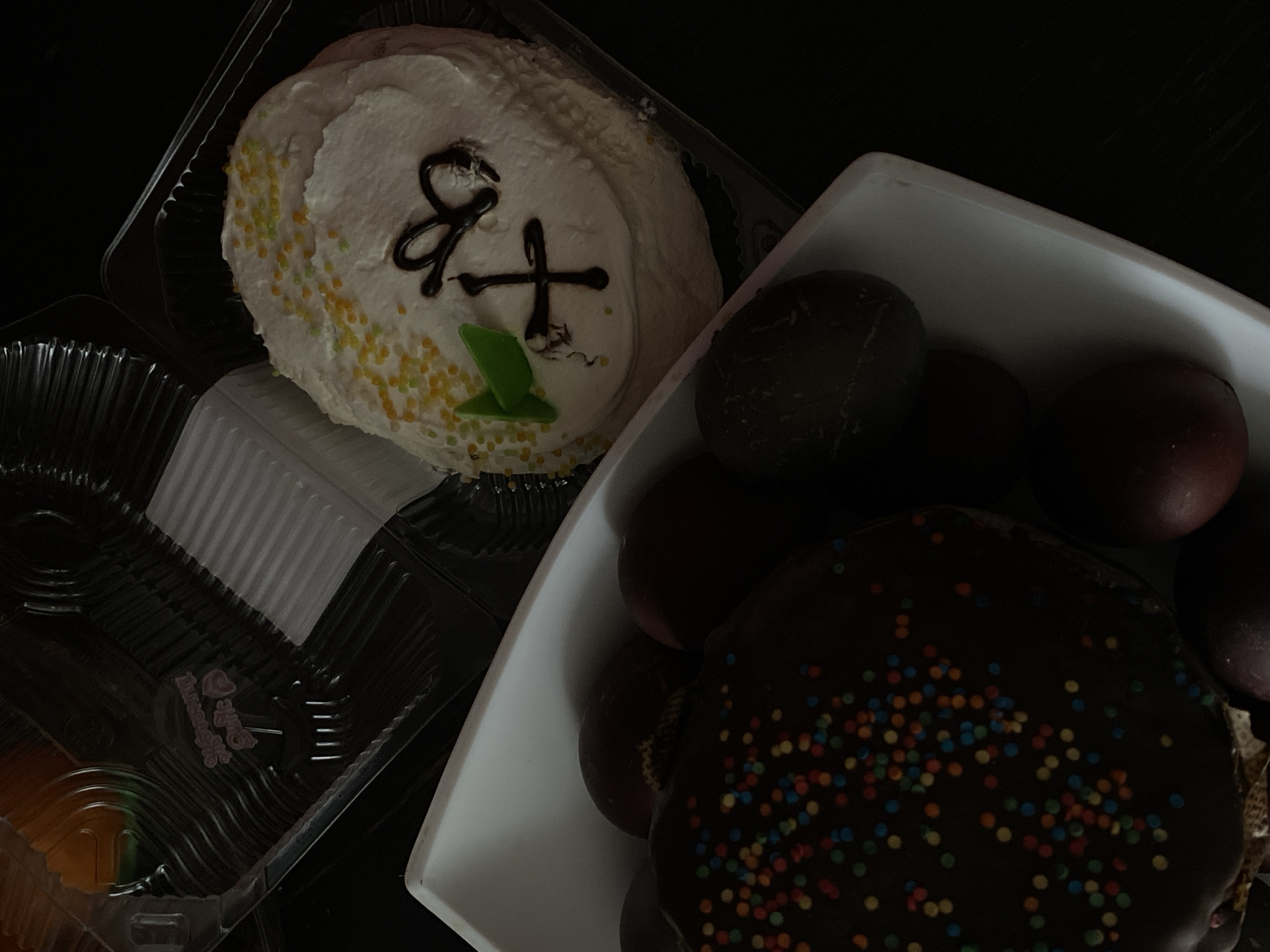 The image shows a dimly lit tabletop scene featuring traditional Easter foods. A white frosted cake with a chocolate design and yellow and green sprinkles sits in a clear plastic container. Beside it, a white rectangular dish holds several colored Easter eggs, with shades ranging from dark green to red-brown. Next to the eggs, a chocolate-covered cake adorned with colorful sprinkles completes the spread. The image's composition suggests a close-up, possibly taken indoors with available light, creating high contrasts and shadows. There's no immediate indication of the location being Chaikovsky, Russia, beyond potential cultural context suggested by the food items.