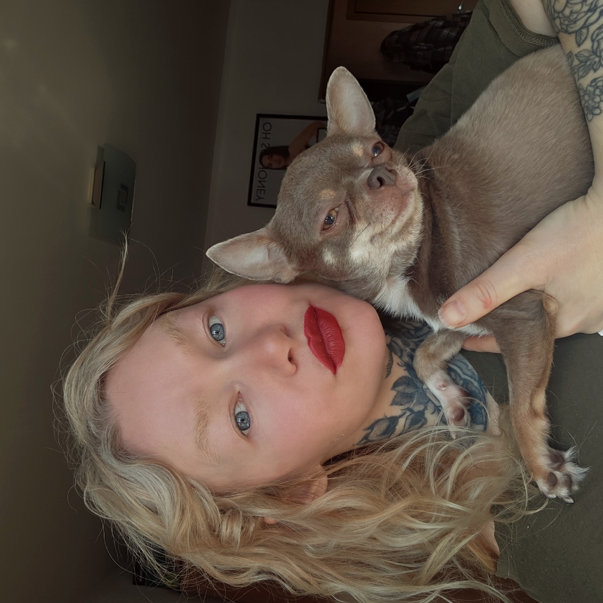 A close-up, rotated portrait features a woman cuddling a small chihuahua dog. The woman has long, wavy blonde hair, fair skin, and bright blue eyes. She wears bold red lipstick, which appears slightly smudged. Intricate floral tattoos are visible on her neck/shoulder and left arm.
She is holding a light brown (fawn) chihuahua with short fur and white markings on its chest and paws. The dog rests its head near hers, looking upwards with soft brown eyes.
The background shows a plain, dark wall. A dark-framed picture hangs on the wall behind the dog; the text on it is mirrored and inverted, but when corrected reads "OH 2 MONEY". A small, light-colored object, possibly a light fixture or small shelf, is visible further back on the wall. The scene is indoors, dimly lit, suggesting a calm and intimate moment.