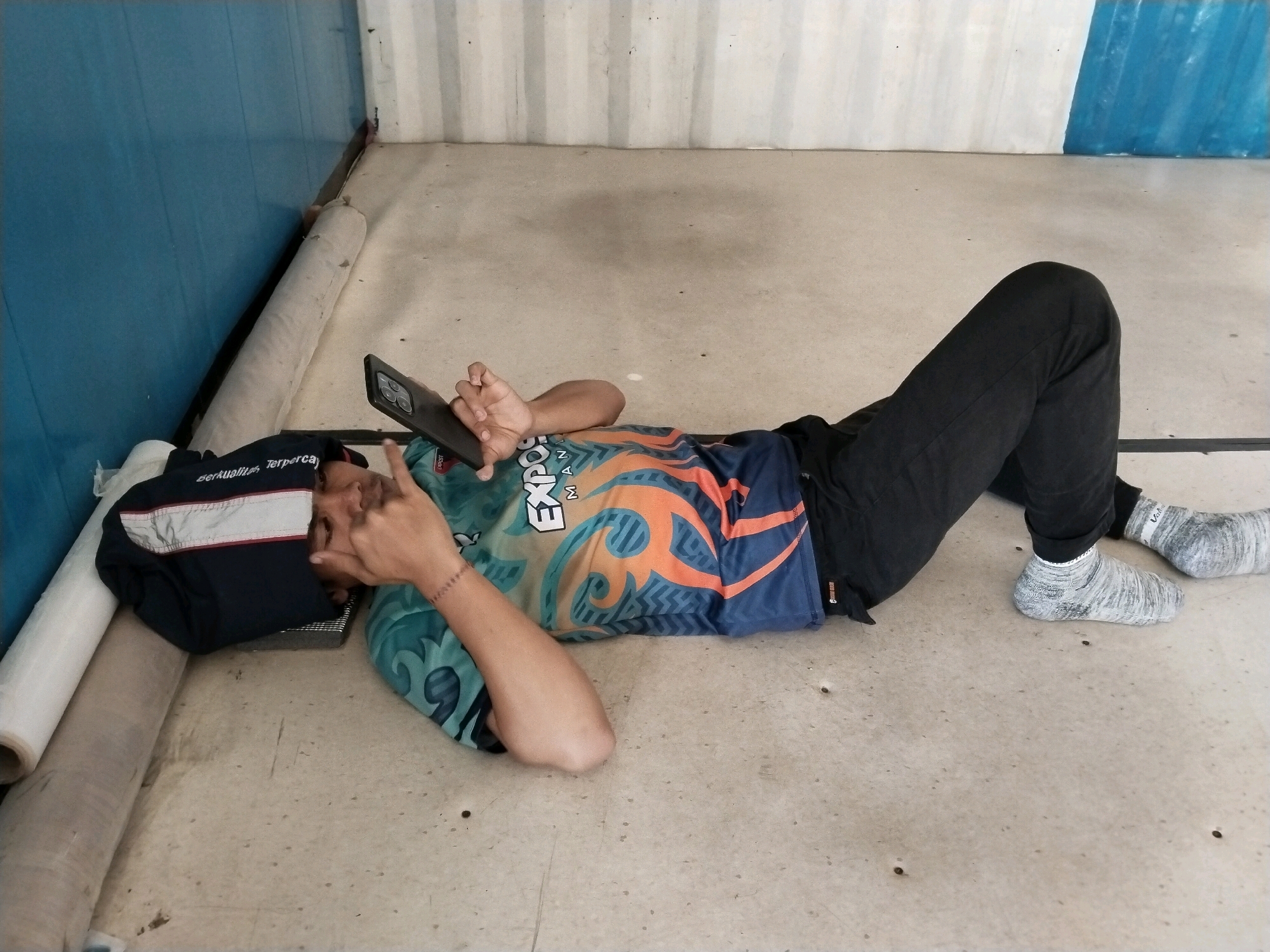 A person is lying on their back on a concrete floor in what appears to be an outdoor or semi-outdoor area, possibly a storage or service area, with a blue corrugated metal wall on the left and a white corrugated metal wall in the background. The person is wearing black pants, patterned socks, and a colorful t-shirt with orange and teal designs and text that appears to read "EXP." and "MAN." They are holding a black smartphone in their hands, looking at it, with their left arm bent and supporting their head. To the left of the person, there is a dark blue jacket or bag with reflective stripes and text that is partially obscured but appears to say "berkualita." A large roll of white material, possibly plastic sheeting or fabric, is also present on the floor. The environment is simple and utilitarian, with visible seams in the wall panels and scattered small holes on the concrete floor.