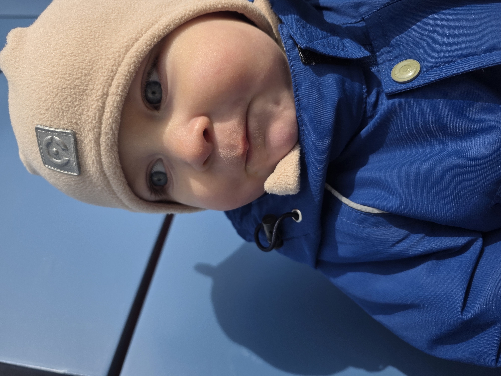 A close-up image depicts a baby situated in the city of Chaikovsky, Russia. The infant, with fair skin and blue eyes, is observed looking towards the upper right of the frame. The baby wears a beige fleece beanie with a silver-colored rectangular emblem featuring a stylized circular logo, and a royal blue puffy winter jacket. A beige collar lining is visible beneath the baby's chin. The jacket features a light-colored drawstring and a black toggle cinch on the left side, and a round, light-colored snap button on the right front closure. The background is a uniform bright blue surface, against which a distinct, dark shadow of an unseen object is cast on the left. A thin, dark horizontal line is visible across the upper left background.