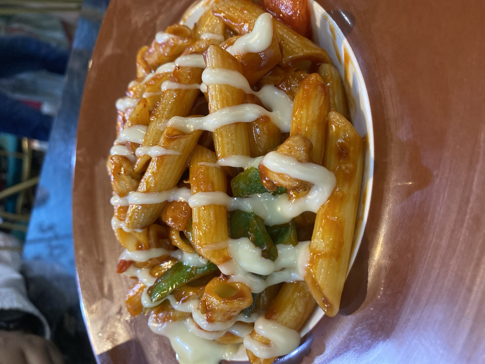 The image shows a close-up of a dish of penne pasta, topped with a red sauce, green peppers, and a white sauce. It is served in a white bowl on a brown surface. Based on available information, the location is Dhaka, Bangladesh. The focus is solely on the presented pasta dish.