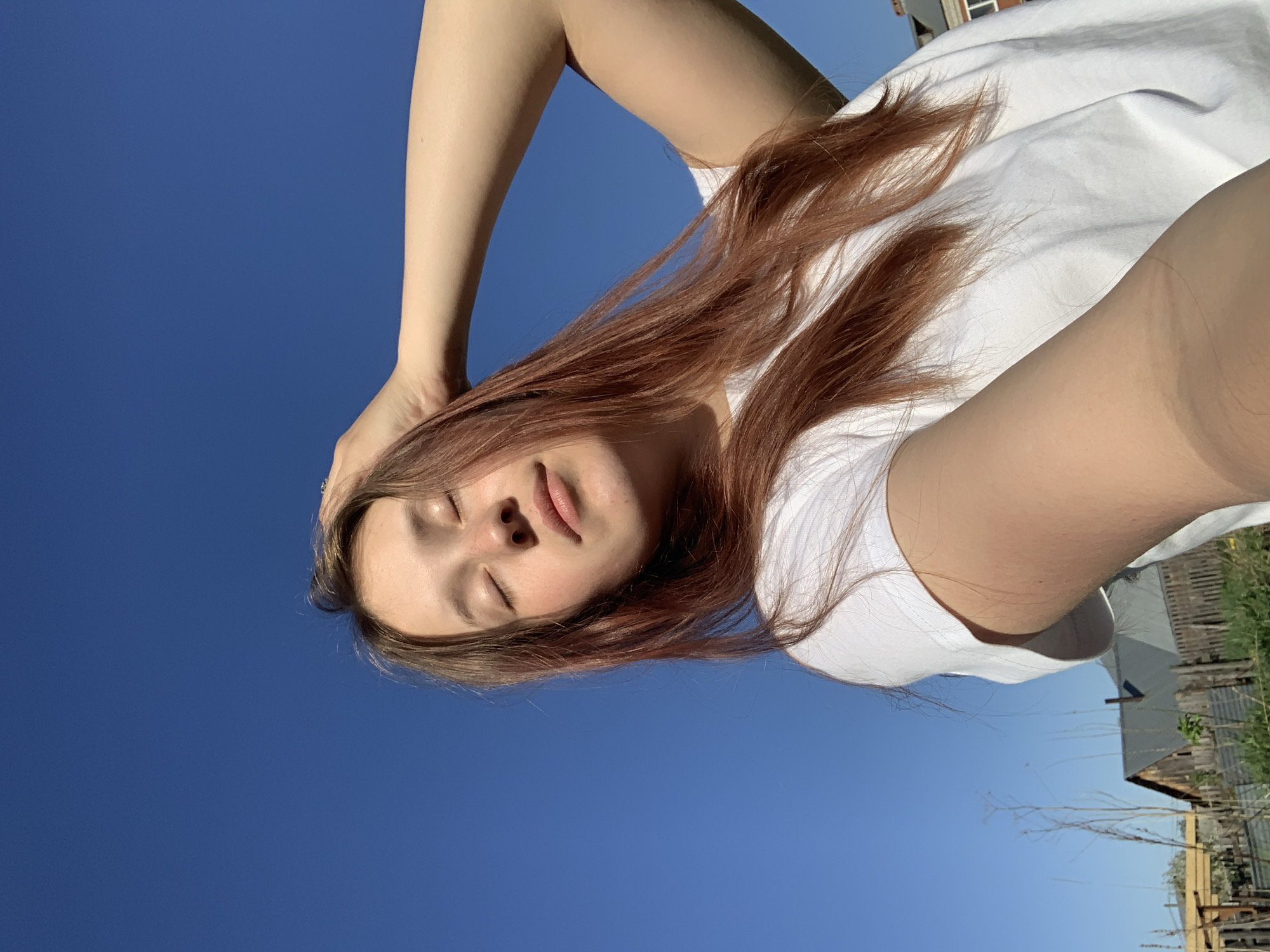 A low-angle shot shows a young woman with light skin and reddish-brown hair, lying on her back. Her eyes are closed and she has a relaxed expression. She wears a white t-shirt. One arm is raised, the hand touching her head. The background is mostly a clear blue sky. In the bottom right corner, there is a glimpse of a rural environment with a building, fencing, and vegetation. Based on the prompt, the location is Фоки, Russia.
