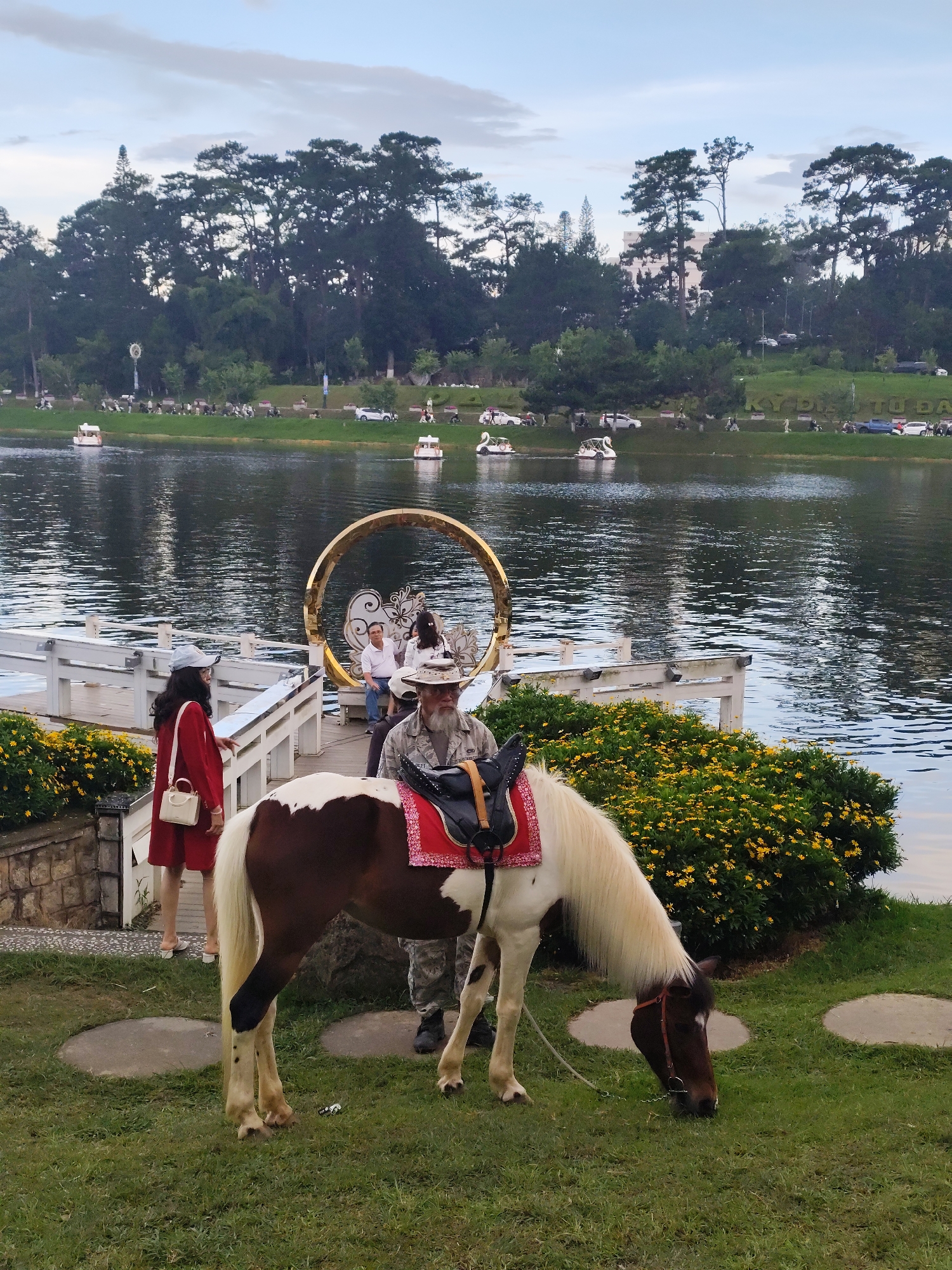 The image depicts a picturesque scene at Xuan Huong Lake in Da Lat, Vietnam. A dappled brown and white pony is grazing on a grassy area in the foreground, with a man in a hat standing next to it. In the background, a woman in a red dress is walking near a white railing, with a golden circular archway adorned with floral motifs behind her. Several swan-shaped paddle boats are visible on the calm waters of the lake, and a line of pine trees forms a dense backdrop along the far shore. The sky above is partly cloudy.