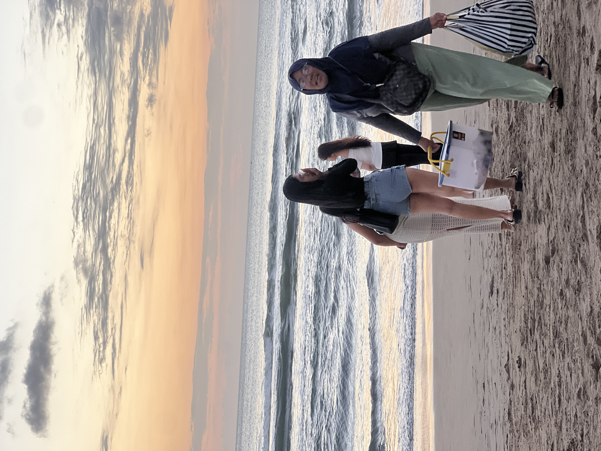 This is a medium shot of a beach in Kuta, Indonesia at sunset. The sky is painted with hues of orange, pink, and yellow, with scattered clouds. The ocean is calm with small waves gently rolling onto the shore. On the sandy beach, four individuals are visible. In the foreground, on the right, a person wearing a dark hijab and green pants is walking towards the viewer, carrying a striped tote bag and a clear plastic bucket with yellow handles. To the left of this person, two women are standing closer to the water. One is wearing a black top and denim shorts, the other a white off-the-shoulder top and black bottoms. Further in the background, another woman is visible with her back to the camera. The sand is damp near the water and drier further up the beach.