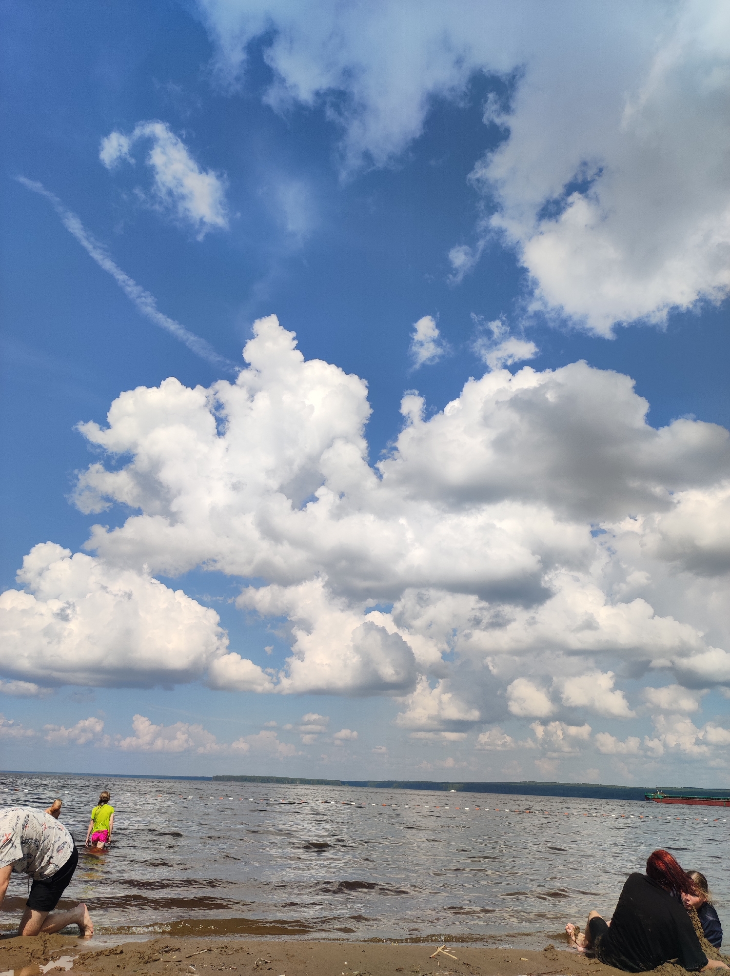 The image captures a scene on the sandy shore of the Kama River in Chaikovsky, Russia, under a bright blue sky filled with fluffy white clouds. The water stretches out, marked by a floating line of buoys. In the foreground, on the beach, three individuals are visible. To the left, a person in a patterned shirt and black shorts is crouching, facing away from the camera. Further into the water, a child in a bright green shirt and pink shorts is partially submerged. On the right side of the beach, a person with dark red hair is sitting, facing the water, with another person partially visible next to them. A large red cargo ship is sailing in the distance on the river. The overall impression is a sunny day at a river beach.