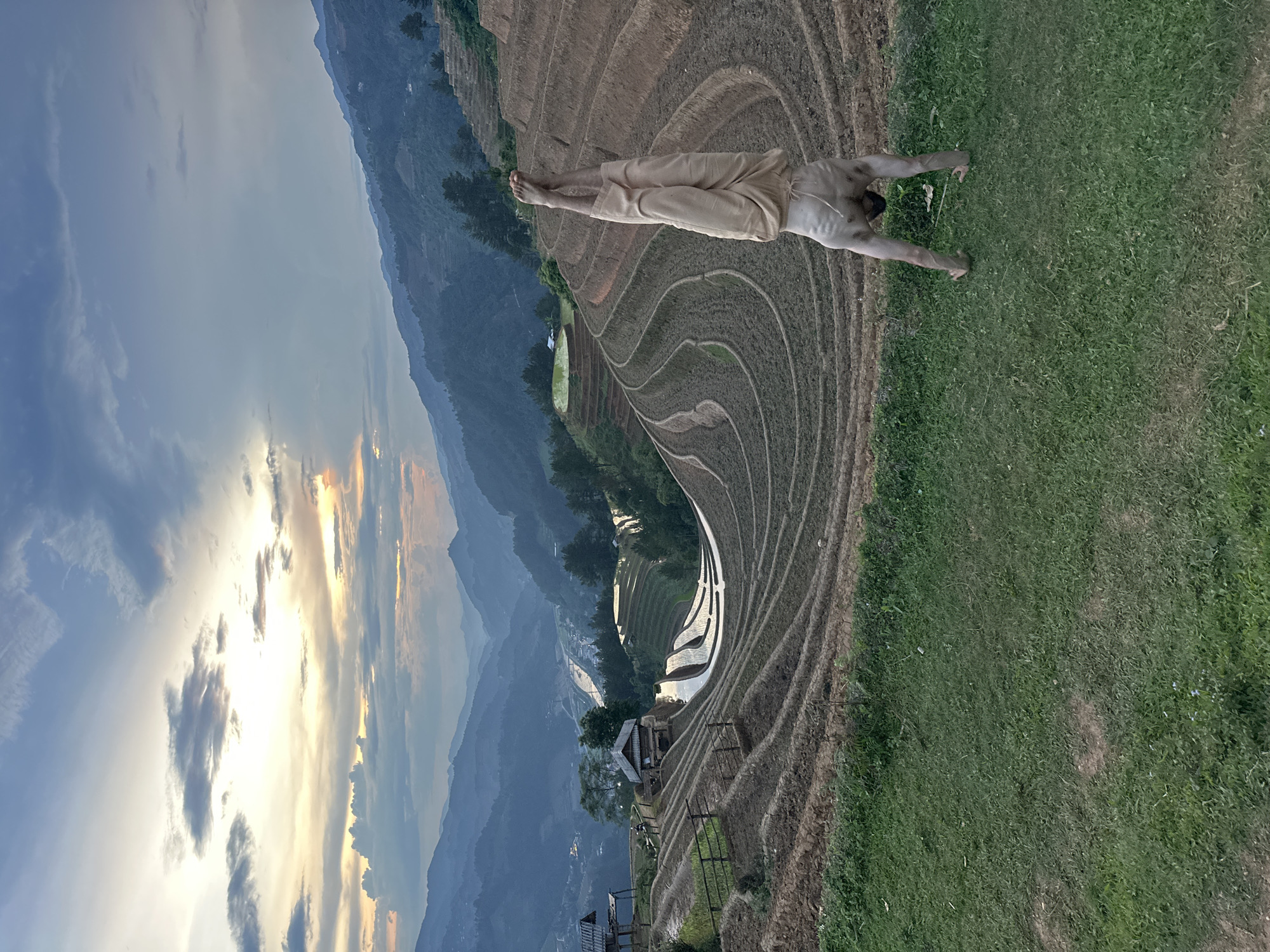 This image captures a serene and expansive landscape of terraced rice fields in Mù Cang Chải, Vietnam, at either sunrise or sunset. In the immediate foreground, a man stands on a patch of green grass, facing away from the viewer. He is shirtless or wearing a light top, with light-colored pants, and has his arms outstretched horizontally, suggesting a moment of peace, contemplation, or appreciation for the view. His back muscles are defined, and he is barefoot.
The dominant feature of the scene is the vast network of meticulously sculpted rice terraces that curve around the undulating hillsides. Some of these terraces in the midground are filled with water, creating reflective surfaces that shimmer under the soft light. Small, traditional structures, possibly huts or houses, are dotted among the terraces, indicating human presence within this agricultural landscape. Beyond the terraces, a range of mountains stretches into the background, fading into a gentle haze.
The sky is a notable element, displaying a beautiful array of colors – soft blues and grays blend with warm oranges and yellows, characteristic of the golden hour. Scattered clouds add texture and depth to the sky. The overall impression is one of tranquility and natural beauty, highlighting the harmony between human cultivation and the dramatic mountain scenery. There is no visible text in the image.