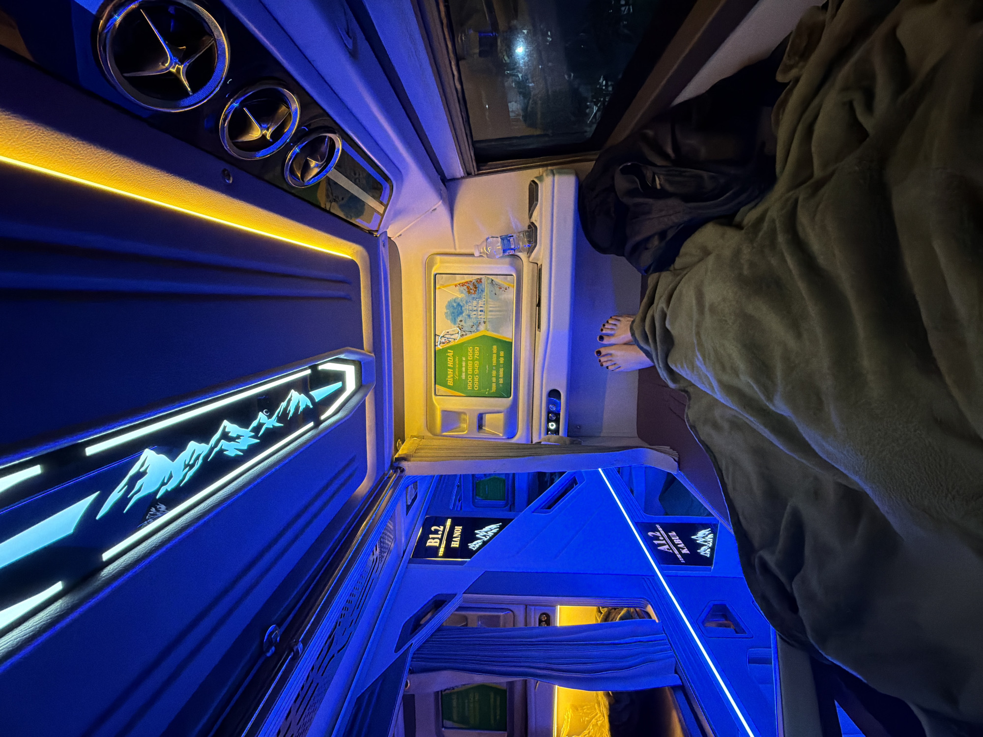 This image captures the interior of a modern sleeper bus or train cabin, likely during a night journey in Vietnam, specifically with context pointing to Đà Nẵng. The perspective is from inside one of the berths, looking down the aisle.
In the foreground, a person's bare feet with painted toenails are visible, resting under a soft, olive-green blanket or comforter. A black bag or backpack is tucked beside their head, suggesting they are lying down and resting.
The cabin is illuminated by vibrant LED lighting. Bright yellow strip lights run along the upper wall of the individual sleeping compartment, providing ambient light. Three circular chrome air vents are visible above this lighting. The main aisle and other parts of the cabin are bathed in striking blue LED lights, including a decorative wall panel featuring a mountain silhouette design.
An information panel with a small landscape drawing and text is affixed to the wall near the person's feet. Visible text on this panel includes "BÌNH HOÀI Limousine," booking hotline numbers "1900 888 666" and "0916 949 789," and route information such as "TOUR HÀ NỘI - THANH HÓA" and "ĐÀ NẴNG = HỘI AN." Further down the aisle, cabin number signs are visible, reading "B1.2 HANOI" and "A1.2 KABUL" (likely a destination or origin). A small plastic water bottle is placed in a holder nearby. Through a window, the darkness of night and some distant artificial lights are visible. The overall ambiance is cozy and conducive to travel and rest.