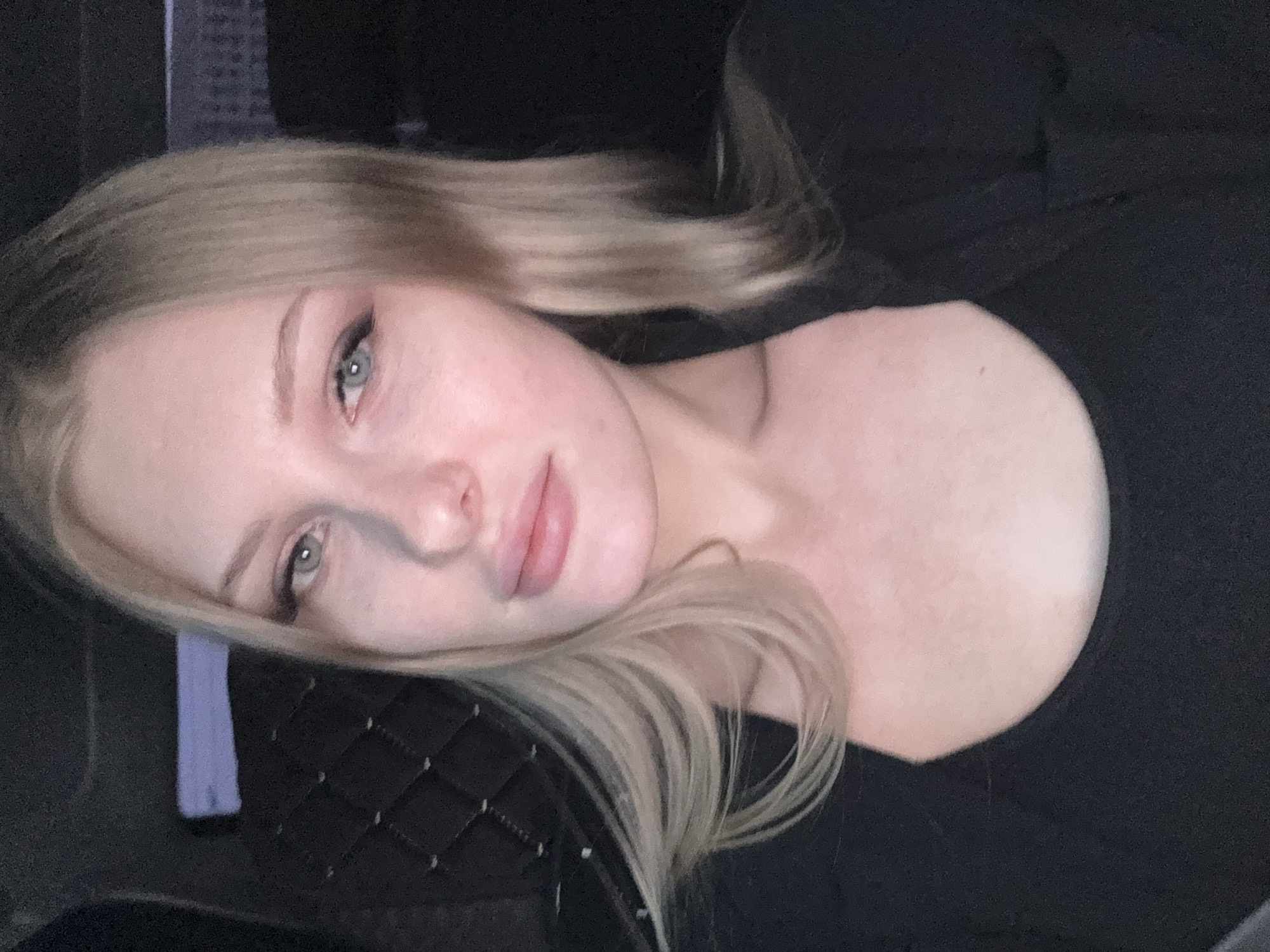 A close-up, rotated portrait features a fair-skinned woman with light blonde hair, wearing a black garment. Her eyes are light-colored, and she has subtle makeup enhancing her features. Part of a quilted black object with a purple strap is visible near her face. The background is dark and indistinct, showing potential features like a screen. The location and its relevance to Perm, Russia, are not discernible from the provided visual data.