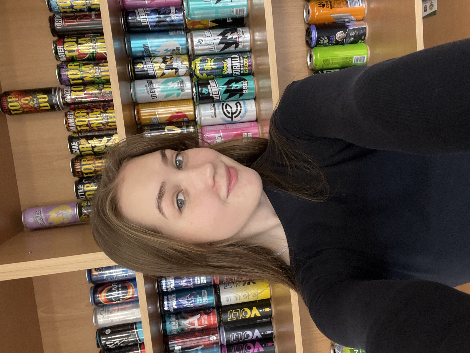 A young woman with light brown hair and light-colored eyes, wearing a black long-sleeved top, is prominently featured on the right side of the frame, looking towards the viewer with a slight, pleasant smile. Her face and upper torso are visible. Behind her, and filling the rest of the image, are numerous rows of assorted energy drink cans stacked horizontally on light-colored wooden shelves.
The setting appears to be an indoor space, possibly a store aisle, a specialized shop, or a collection display, given the sheer volume and variety of energy drink brands. The lighting is bright and even, consistent with an indoor environment.
Numerous energy drink brands and flavors are visible on the cans, including "TOP" (with flavors like Watermelon, Razzle, Mango, Cake, Lemonade, DU, Storm), "LITE ENERGY" (Original, Mango Berry Coconut, Blue Raspberry, Classic, Malina Crush), "GORILLA Green," "SOLAR POWER," "Adrenaline Rush" (Game Fuel, Limited Edition), and various "VOLT" cans. Other visible text includes "ENERGY," "ORIGINAL," "LIMITED EDITION," "GAME FUEL," "MAXB," "ENRGT," "ENE," "Jaguar," "F," and "24 МИЛЛИОНЕ" (24 million). The woman seems to be taking a selfie or being photographed in front of this extensive display of beverages in Chaikovsky, Russia.