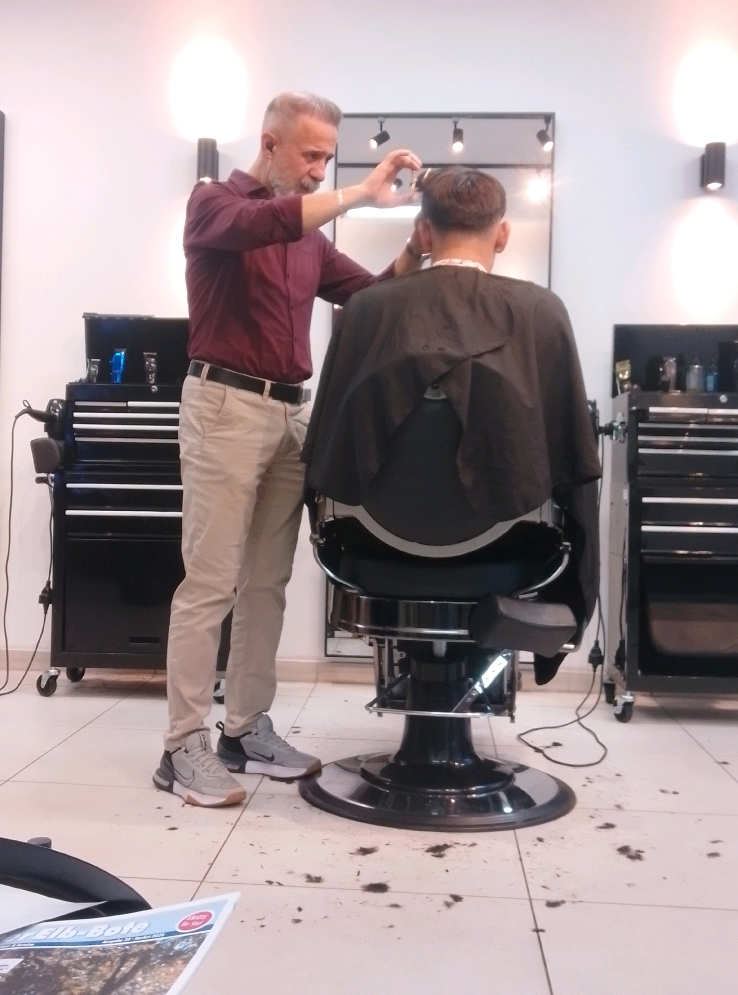 In a barbershop, a male barber with graying hair and a beard, wearing a maroon button-down shirt and tan pants, is cutting the hair of a male customer sitting in a black barber chair, who is draped in a dark cape. The barber is holding scissors in his right hand and appears to be using his left hand to section the customer's hair. The floor is tiled and scattered with cut hair. To the left of the barber is a black rolling tool chest with drawers, containing barbering equipment. To the right of the barber chair is another black rolling tool chest. The background shows white walls with two wall-mounted lights, a mirror reflecting the scene, and more barbering equipment on shelves. A magazine is visible on the floor in the foreground. The scene is set in a modern, well-lit barbershop. This barbershop is located at Hans-Böckler-Straße 26, 22880 Wedel, Germany.