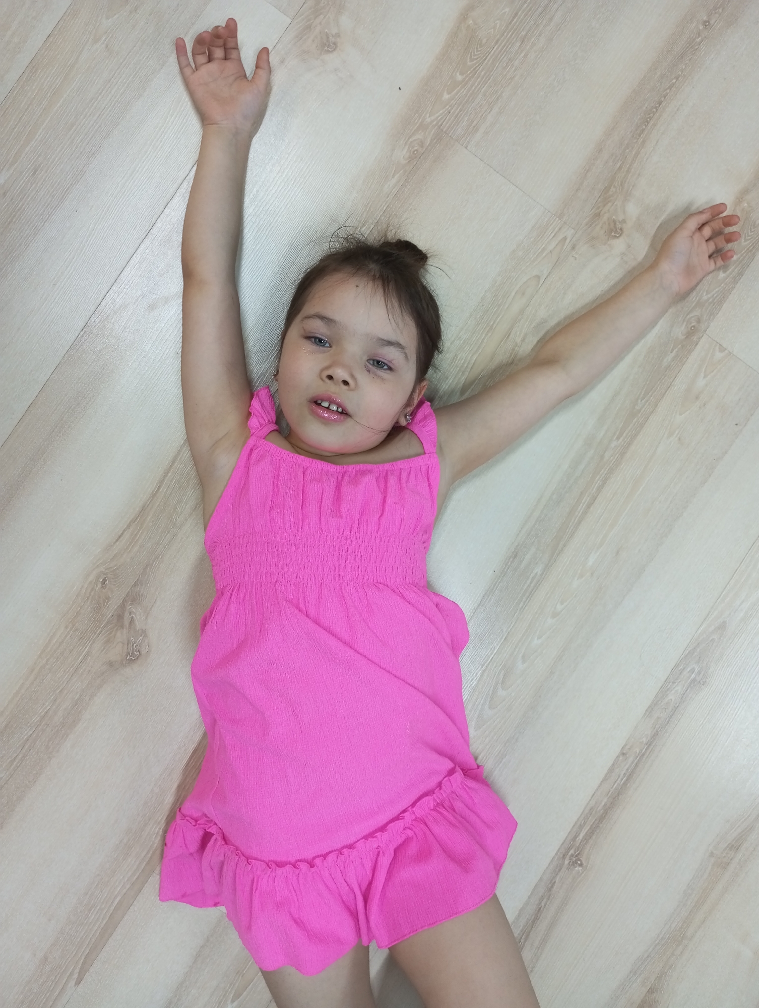 A young girl wearing a bright pink dress is lying on her back on a light wood-look laminate floor. Her arms are outstretched to the sides and slightly above her head. She has brown hair pulled back, fair skin, and light-colored eyes. The background is out of focus. The location is not discernable as Pervomaysky, Russia based on the image content alone.