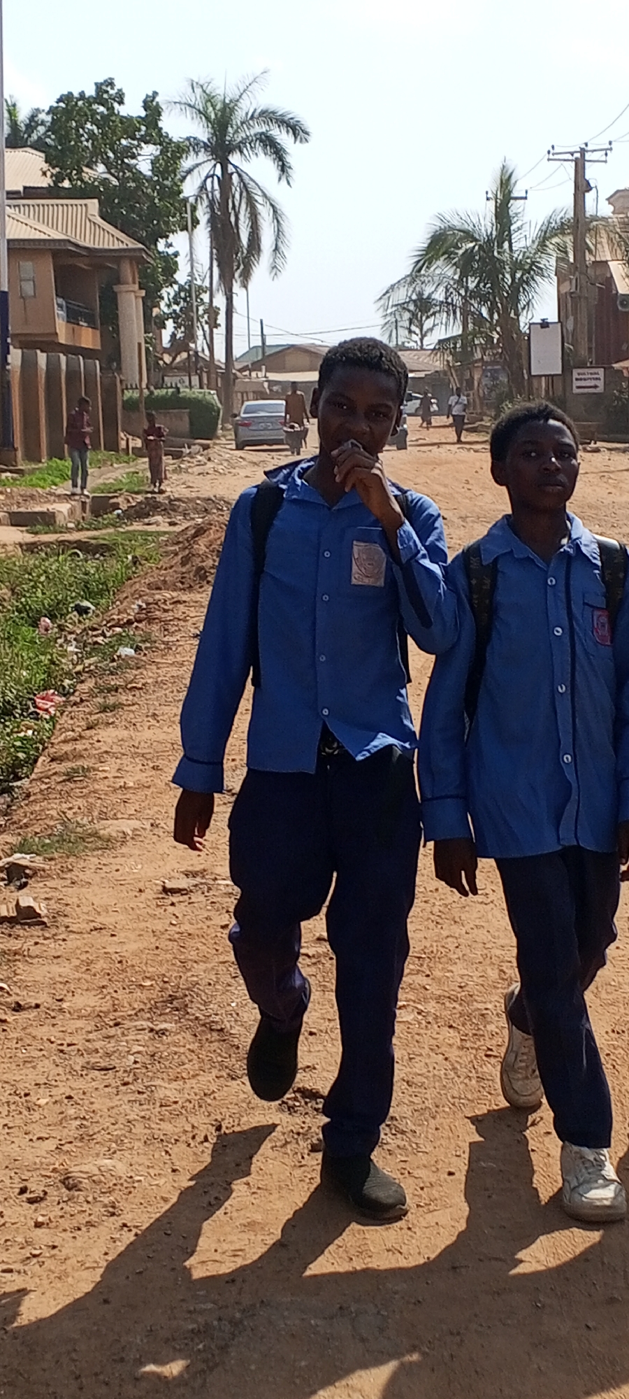 Two young Black male students in blue school uniforms with backpacks walk on a dirt road in Sabon Tasha, Nigeria. The student on the left has his hand to his mouth. Buildings, palm trees, and a car are visible in the background. Two children and people walking are also present in the background.