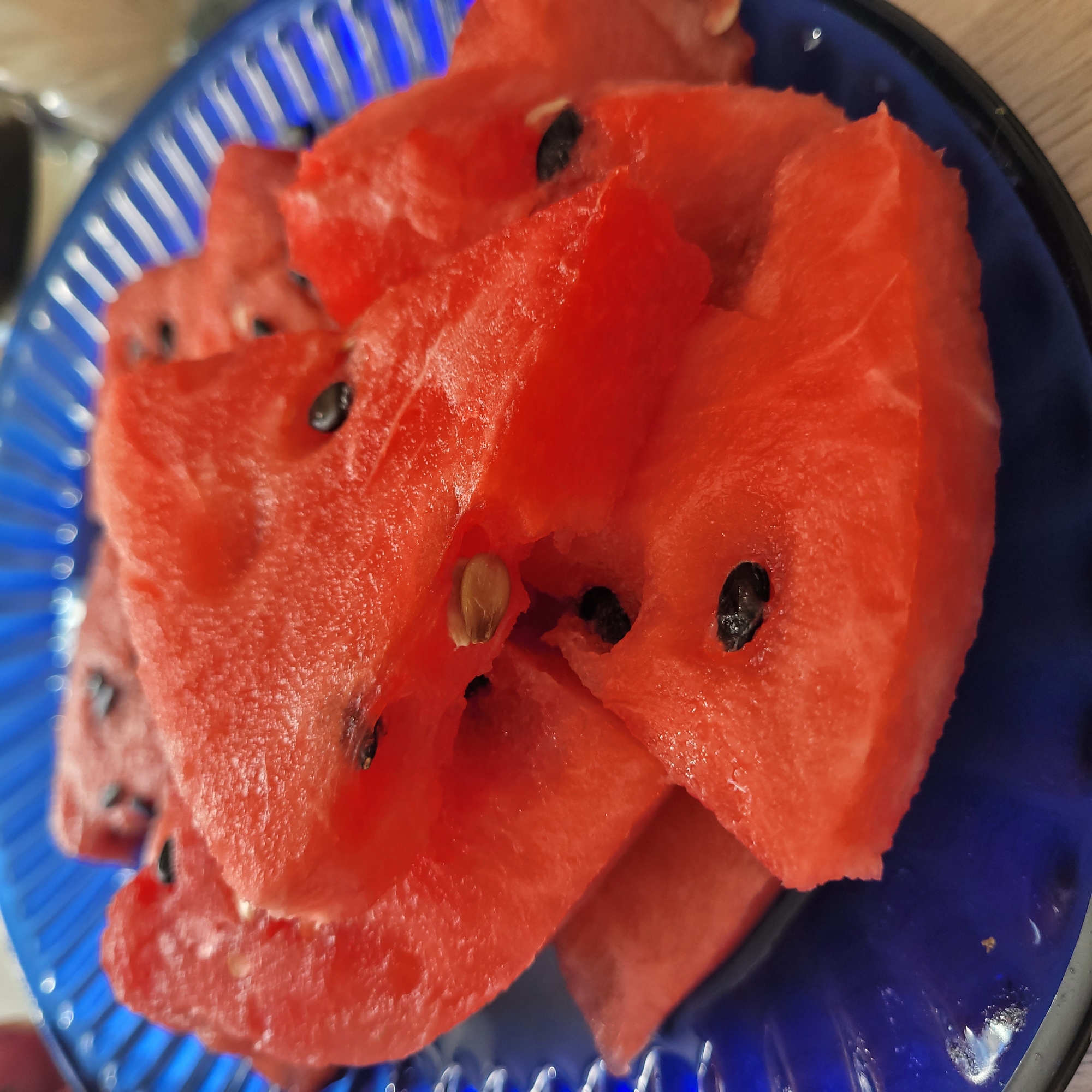 A close-up shot shows several slices of ripe watermelon on a blue, ribbed glass plate. The watermelon slices are a vibrant red and some contain dark seeds. The plate is positioned on a light-colored wooden surface. The background is blurred, but hints of bright lights suggest an indoor setting. There is no indication of the specific location within Jūrmala, Latvia.