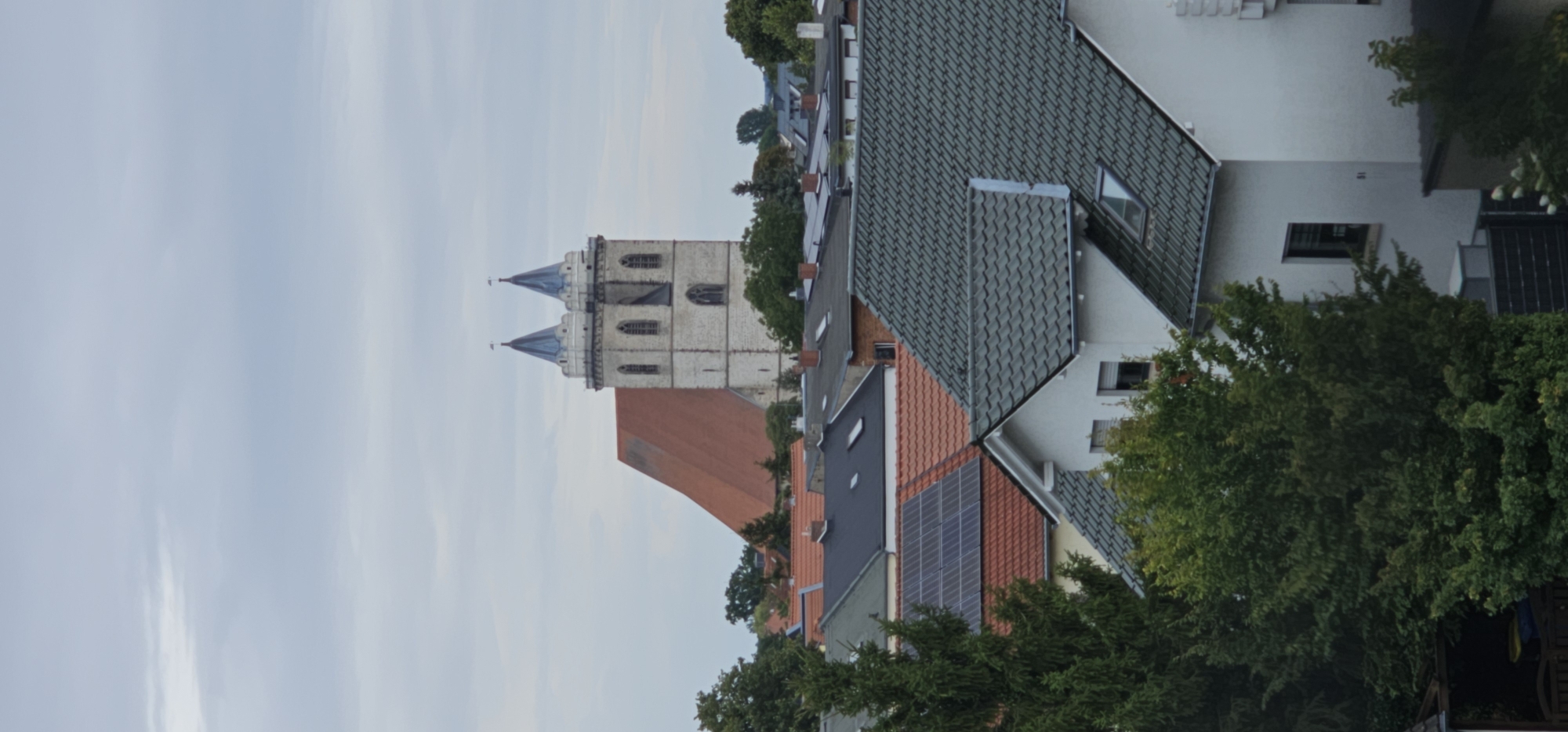 The image shows an overhead view of a city with residential buildings and a large church with two spires in the background. The sky is overcast. The foreground is dominated by the roofs of several houses, with some trees visible on the left side of the image. Solar panels are installed on the roof of one house. The church has a large red roof and light-colored stone walls. The spires are pointed and blue.