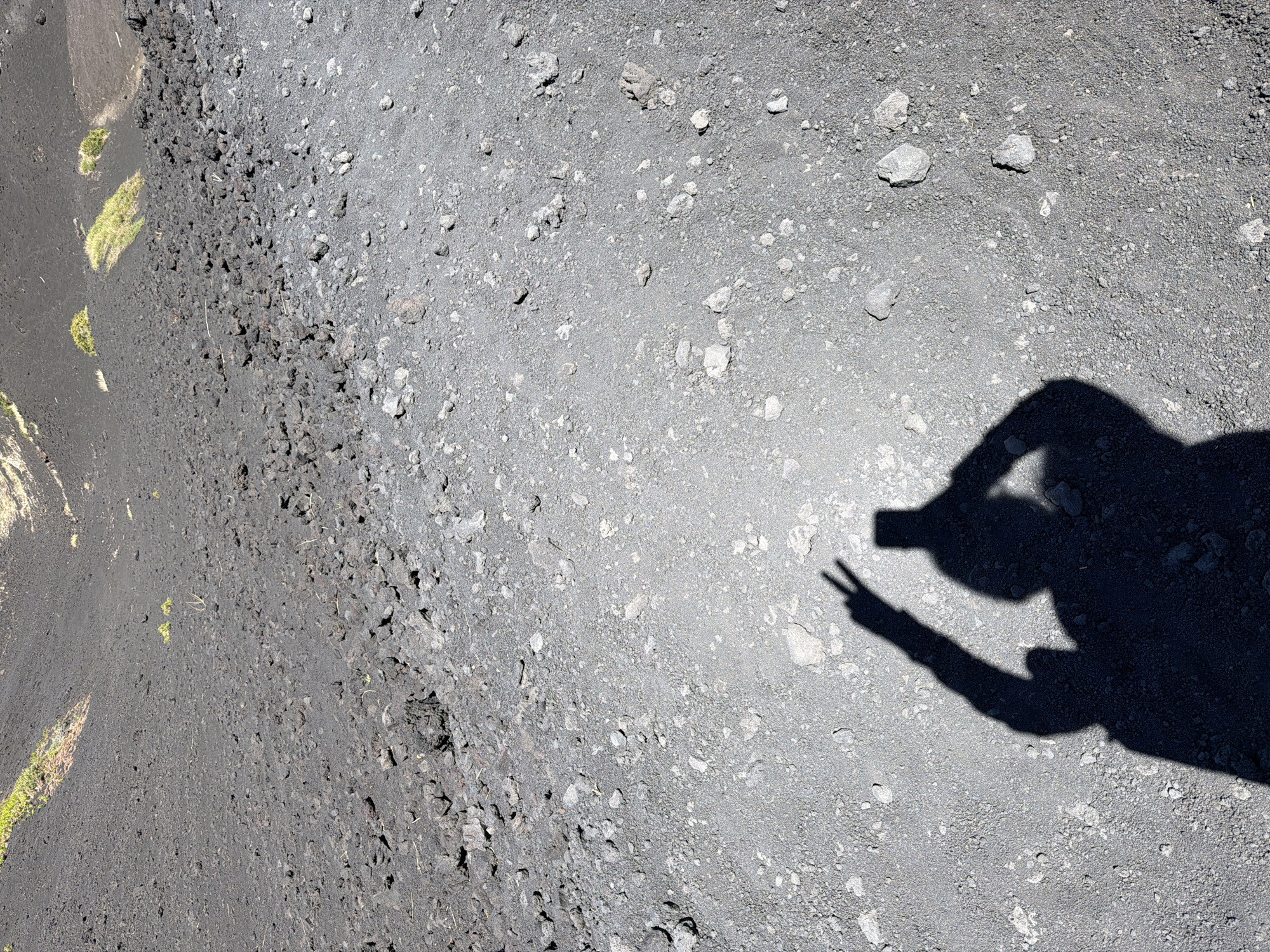 The image displays a shadow of a person on the rocky, volcanic terrain of Mount Etna, near Linguaglossa, Italy. The shadow, cast by the sunlight, is in the lower right quadrant of the image, showing a figure holding a smartphone and making a peace sign with their left hand. The terrain is composed of dark volcanic ash and scattered rocks of various sizes. In the background, patches of green vegetation are visible on the slope of the volcano. The lighting suggests it is a clear day with strong sunlight.