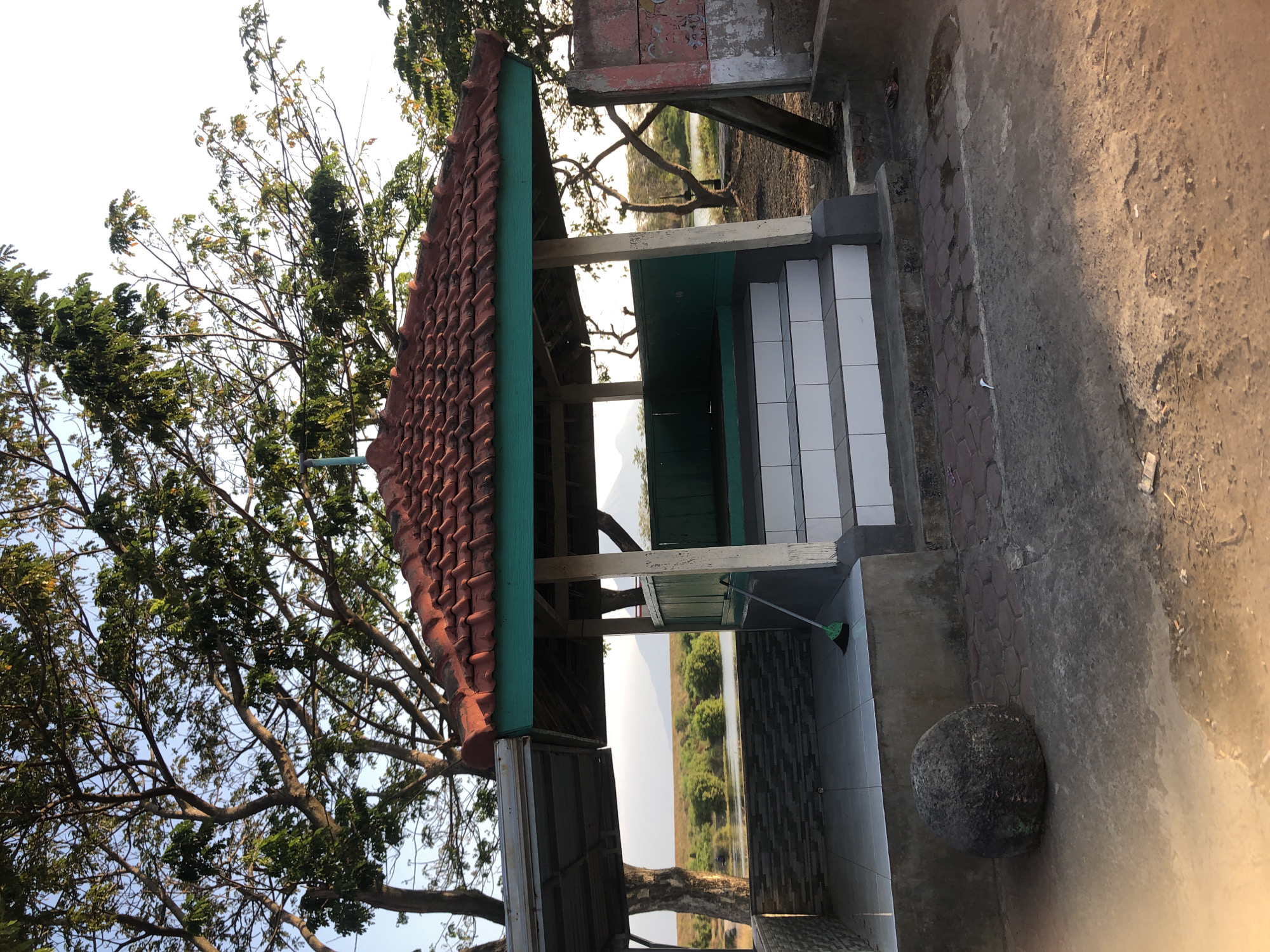 The image shows a gazebo with a red tiled roof and green trim, located outdoors in Kalitengah, Indonesia. The gazebo is elevated with white-tiled steps leading up to it. Inside the gazebo, there appears to be a raised platform, also tiled. A green broom leans against the structure. To the left of the gazebo, there's a covered area with a metal roof. In the foreground, a large, rough, dark gray boulder sits on a brick-patterned surface. Behind the gazebo, a body of water is visible, with trees and distant mountains under a clear sky. The overall scene suggests a park or recreational area.