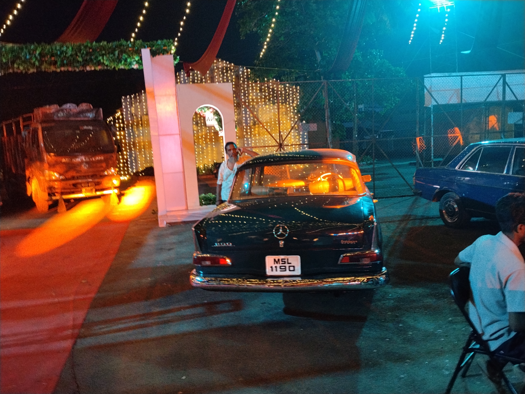 This nocturnal image captures a festive outdoor event, likely in Kalamassery, India, characterized by a dynamic interplay of artificial lighting.
In the middle ground, a vintage, dark-colored Mercedes-Benz sedan is prominently featured, viewed from its rear. The car's chrome bumper reflects ambient light, and its license plate displays "MSL 1190". Text on the trunk appears to indicate a "220 S" model. A man in a white shirt stands beside the car, near a white, light-adorned archway, looking towards the viewer.
To the left, a truck, possibly an Ashok Leyland (based on visible text on its front), is parked with its bright headlights casting a strong orange glow onto the ground. Overhead, red fabric drapes and green foliage garlands are strung with numerous warm fairy lights, creating a celebratory atmosphere. These vertical strings of lights also decorate structures behind the vehicles.
The right side of the scene is illuminated by a distinct cool blue light from an overhead source. Here, a blue sedan is parked behind a chain-link fence. In the immediate foreground on the right, the back and side of a person seated on a chair are visible, wearing a light-colored shirt. The ground beneath exhibits a striking contrast between the orange light spill from the truck and the cool blue ambient light.
The overall setting, with decorative lights, vintage vehicles, and people gathered, suggests a special occasion, perhaps a car display, wedding, or a community event.