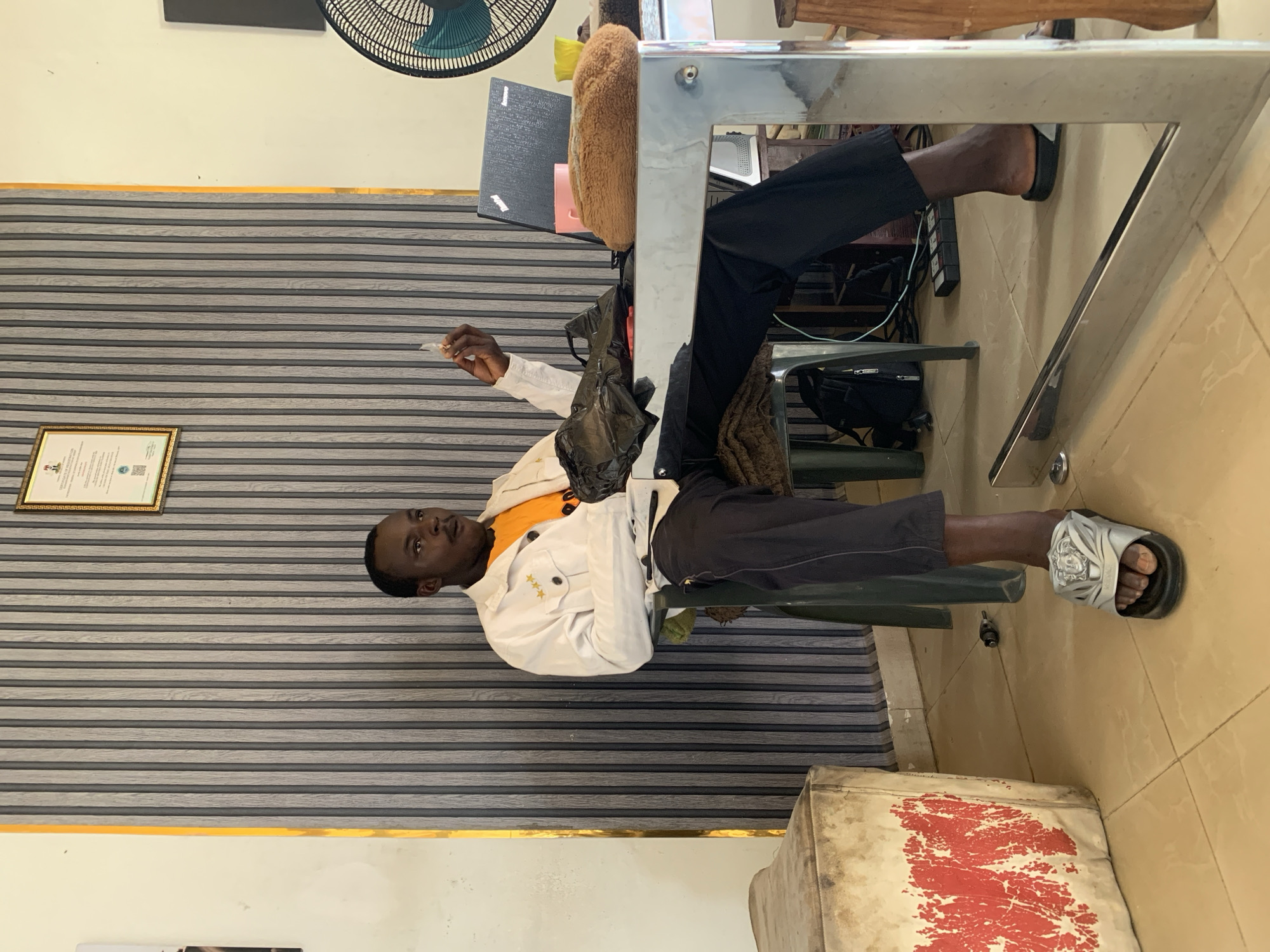 The vertically oriented image captures a young Black man seated indoors, likely in an office or commercial space in Jos, Nigeria. He is positioned on the right side of a reflective, silver-toned metal desk or table.
The man wears a white long-sleeved jacket adorned with three golden star-shaped embellishments on the left chest, an orange t-shirt, and dark trousers. He has dark sandals featuring a circular emblem on his feet. In his right hand, he holds a small, white, folded item, possibly paper or a snack. A black plastic bag rests near his left arm on the table. He looks towards the right side of the frame with a neutral expression.
The background wall features horizontal gray and dark gray stripes, topped by a golden strip and a plain light-colored wall. On the striped section, a framed certificate is visible, bearing text such as "FEDERAL REPUBLIC OF NIGERIA," "REGISTRATION CERTIFICATE," and "CAC," along with a QR code. A wall-mounted black fan is seen in the upper left corner.
Beneath the desk, a power strip and a dark bag are visible. To the right, on the tiled floor, rests a worn, light-colored stool or box with red, abstract markings. A brown, fur-like object is placed under the desk. The scene suggests a functional, possibly modest, indoor environment.