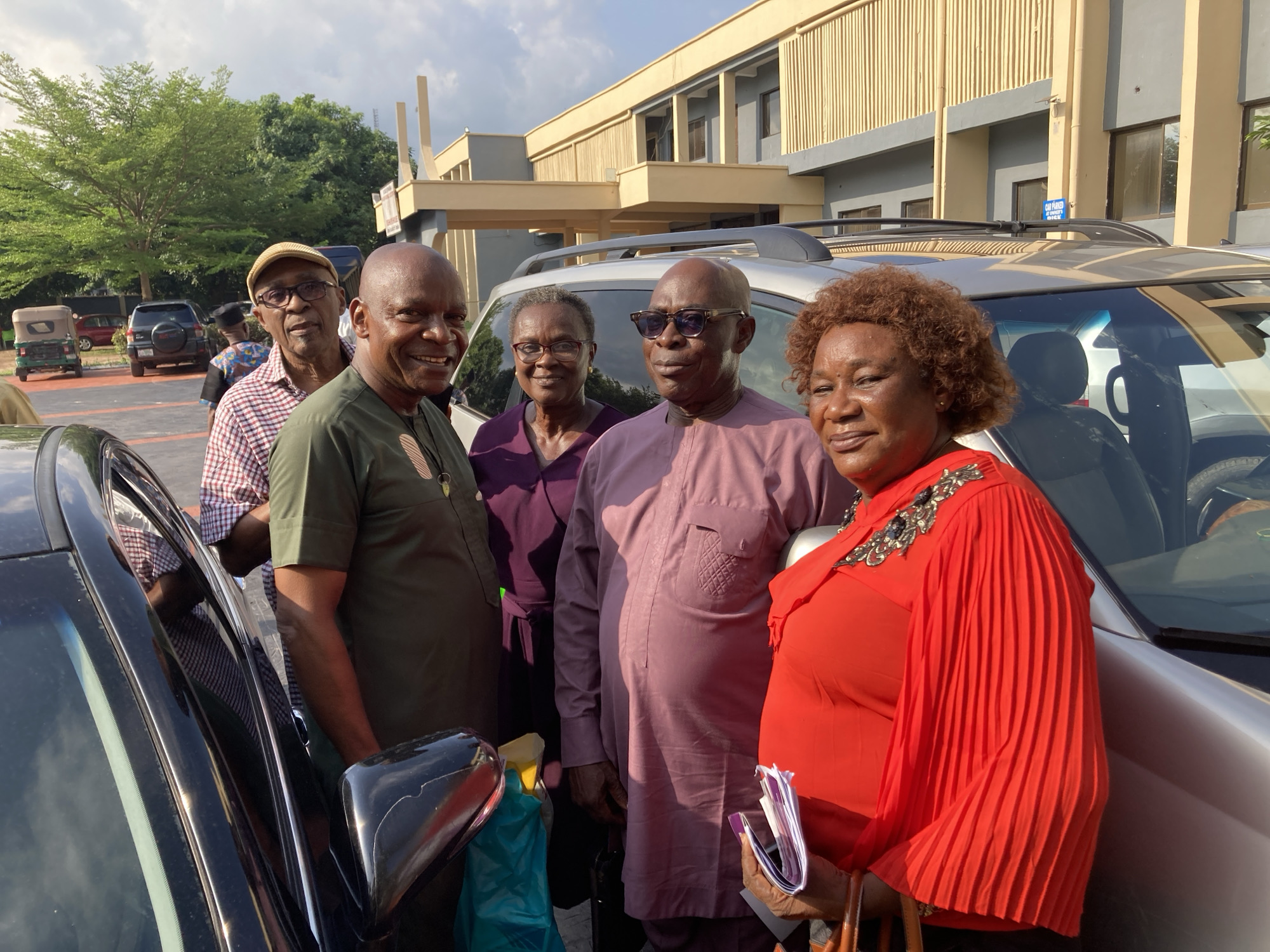 The image shows a group of five adults, appearing to be middle-aged to elderly, standing outdoors in what looks like a paved parking area or forecourt in Umuahia, Nigeria. All five individuals are of African descent and are smiling or looking pleasantly towards the viewer, conveying a friendly and positive mood.
From left to right, partially obscured by a black car, a man in a plaid shirt and a beige cap is visible. Next to him, a man in a dark green traditional top with embroidery smiles broadly. Behind him and to the right, a woman in a purple top and glasses offers a gentle smile. Adjacent to her is another man wearing a lavender traditional top and dark sunglasses. On the far right, a woman with curly auburn hair is dressed in a vibrant red pleated top featuring intricate embellishments on the collar, holding a purple folded booklet.
Two cars are prominent: a black car on the left and a silver SUV behind and to the right of the group. A turquoise bag and a yellow item are visible on the ground near the black car. In the background, a "keke napep" (tricycle taxi) and another SUV are parked further down the paved area.
The background features a modern building with a facade of beige and grey panels, suggesting an office or institutional structure. Green trees are visible beyond the building and parking area. The bright lighting and distinct shadows indicate it is daytime, likely late morning or afternoon, under clear weather. A partially visible blue sign on the building reads "CAR PARKED...". The scene suggests a casual gathering or group photo opportunity.