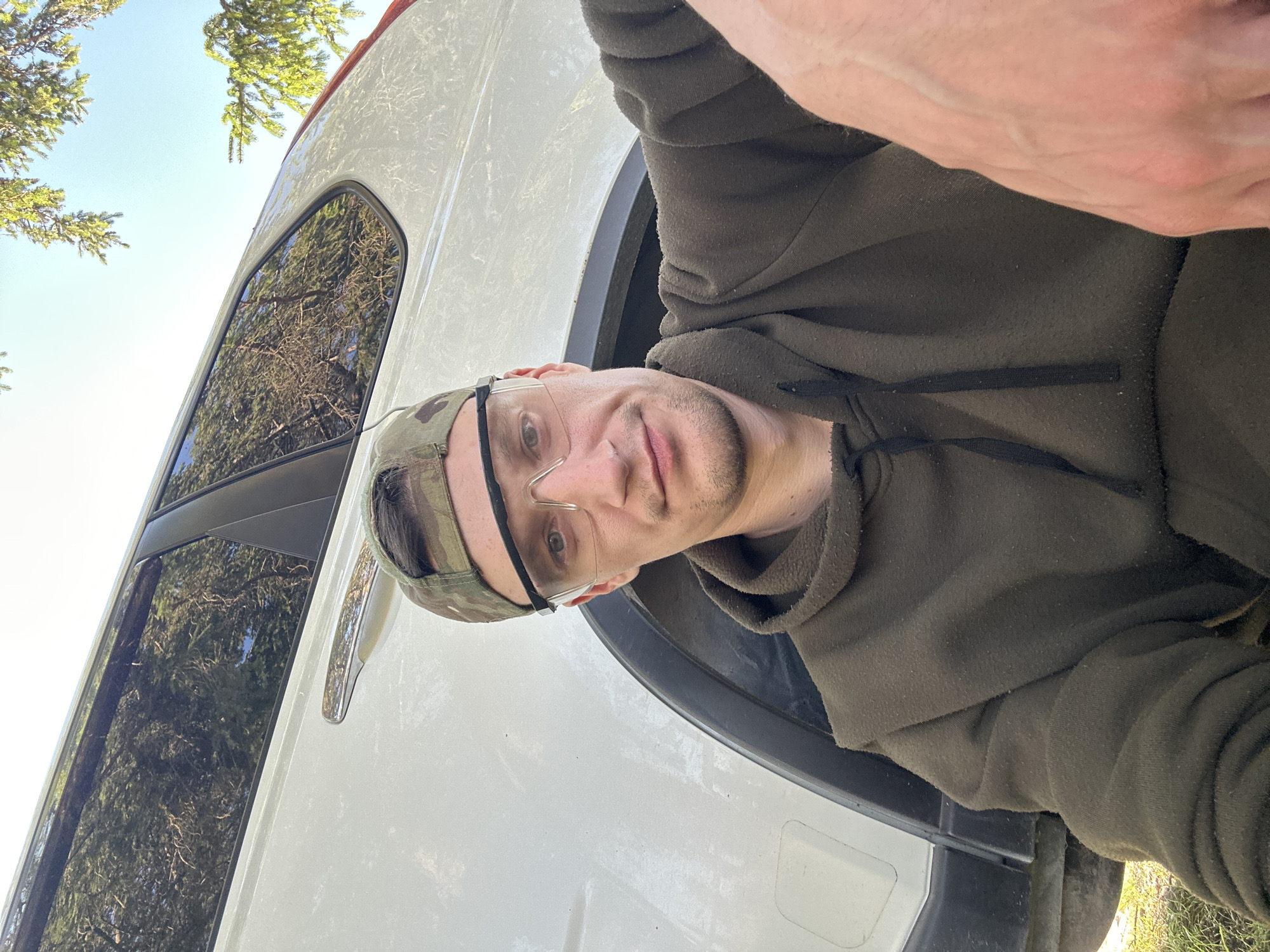 A selfie is taken from a low angle by a man wearing a camouflage baseball cap and clear safety glasses. He is dressed in a dark olive hooded sweatshirt. The man is positioned in front of a white SUV, with the lower portion of the vehicle and a patch of grass visible in the foreground. Trees are reflected in the tinted windows of the SUV and their branches extend into the top of the frame. The sky above is clear and blue. The man is looking directly at the camera with a neutral expression. His right hand is visible in the lower right corner of the image, with veins prominent. The scene appears to be outdoors, possibly in a rural or suburban setting given the presence of the SUV and greenery. The location of Makiv, Ukraine is not discernible from the visual information in the image.