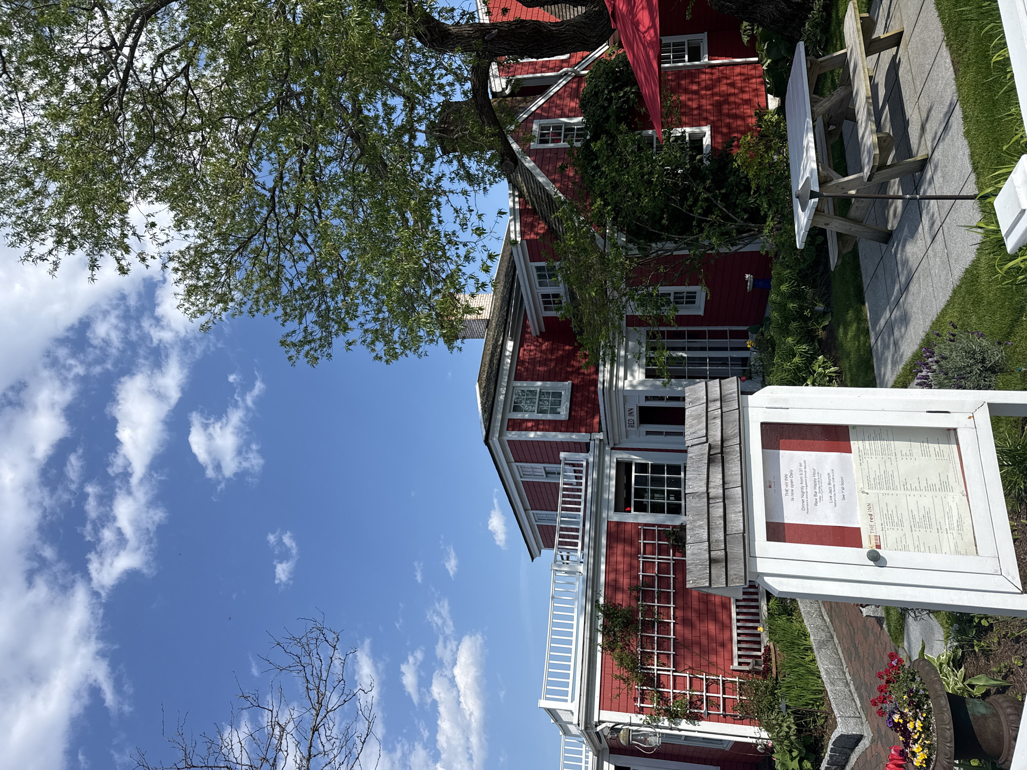 The image shows The Red Inn, located in Provincetown, USA. A red wooden building with white trim and multiple windows stands prominently. A tree with green leaves occupies the upper-left corner, partially obscuring the building. A white picket fence is visible on the building's balcony. A sign displays the restaurant's name, 'THE red INN,' and announces its opening hours. A white picnic table sits on a stone patio next to the building. The sky is blue with scattered white clouds.