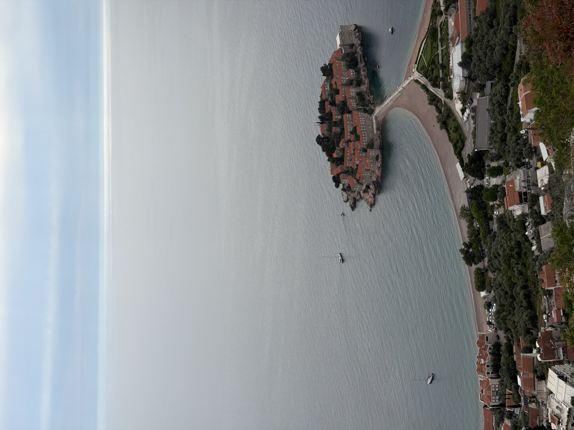 The image presents an aerial view of Sveti Stefan, Montenegro, specifically focusing on the island resort connected to the mainland by a narrow isthmus. The island features a dense cluster of red-roofed buildings amidst greenery, surrounded by the Adriatic Sea. The water is textured with subtle ripples, suggesting a calm surface. To the right of the island, the coastline of Montenegro extends, with buildings and lush vegetation lining the shore. Several small boats are scattered on the water. The sky is visible on the left, fading from blue to a hazy grey, indicating atmospheric conditions like fog or distant haze.