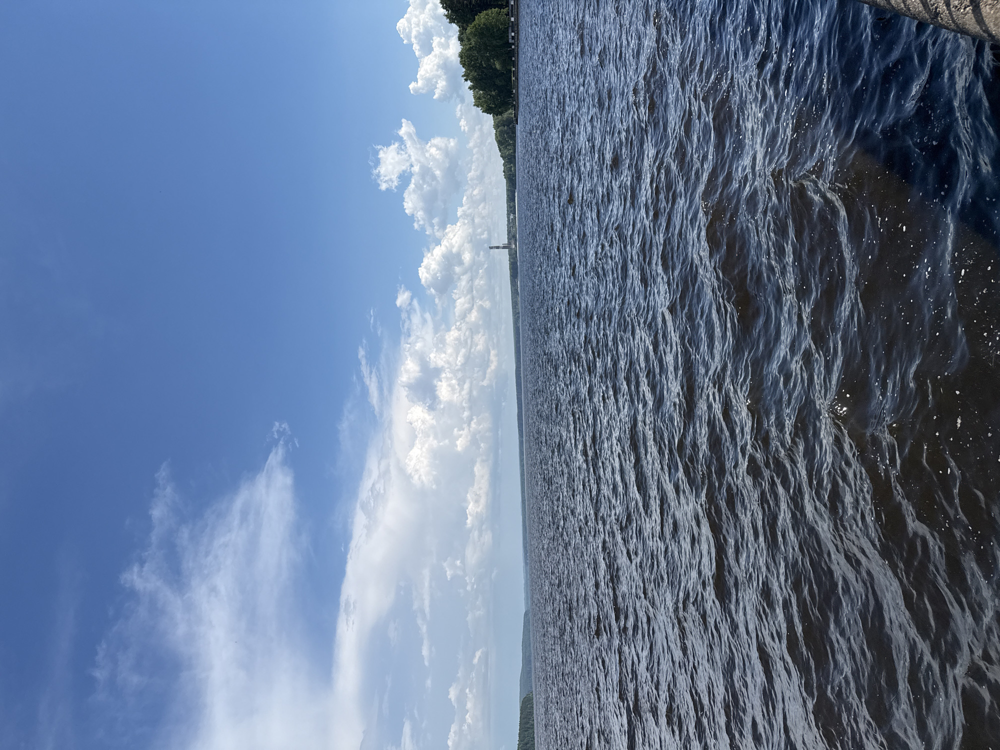 A wide shot captures a vast body of dark, rippling water under a bright blue sky dotted with scattered white clouds. The horizon is marked by a distant, tree-lined shore, with a tall, slender structure resembling a radio tower visible on the left side of the frame. The water takes up roughly two-thirds of the image, with the sky occupying the remaining upper third. A hint of a railing or edge of a structure is visible in the lower right corner.
