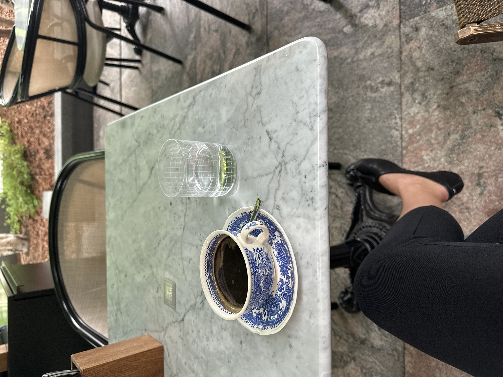A high-angle, medium shot captures a person's legs and feet, clad in black trousers and black ballet flats, at a marble-topped table in what appears to be an outdoor cafe setting. The table surface displays a white marble with grey veining, on which rests a blue and white patterned ceramic cup filled with dark coffee, accompanied by its matching saucer and a small silver spoon. To the right of the coffee cup is a clear glass with a grid pattern, containing a small amount of water with a green sprig at the bottom. In the background, blurred chairs with woven seats and metal frames are visible, as well as greenery and tree trunks, suggesting an outdoor urban environment. The flooring beneath the table is a patterned grey and pink stone. The scene does not provide any specific visual cues to indicate its location within Saint Petersburg, Russia.