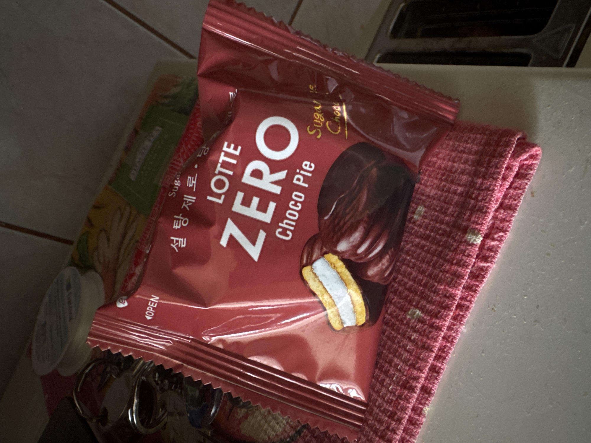 A red package of LOTTE ZERO Choco Pie is displayed on a pink textured cloth. The package is partially open, revealing a drawing of a Choco Pie with a cross-section showing the cake layers and filling, and several chocolate-coated pies. The text on the package includes "OPEN", "LOTTE ZERO", and "Choco Pie". To the left of the package, a set of keys is visible. The background is dimly lit and appears to be a countertop or table surface. The image does not provide any information about the city of Kholmsk, Russia, or any specific environmental details that could be used for forensic analysis related to that location.