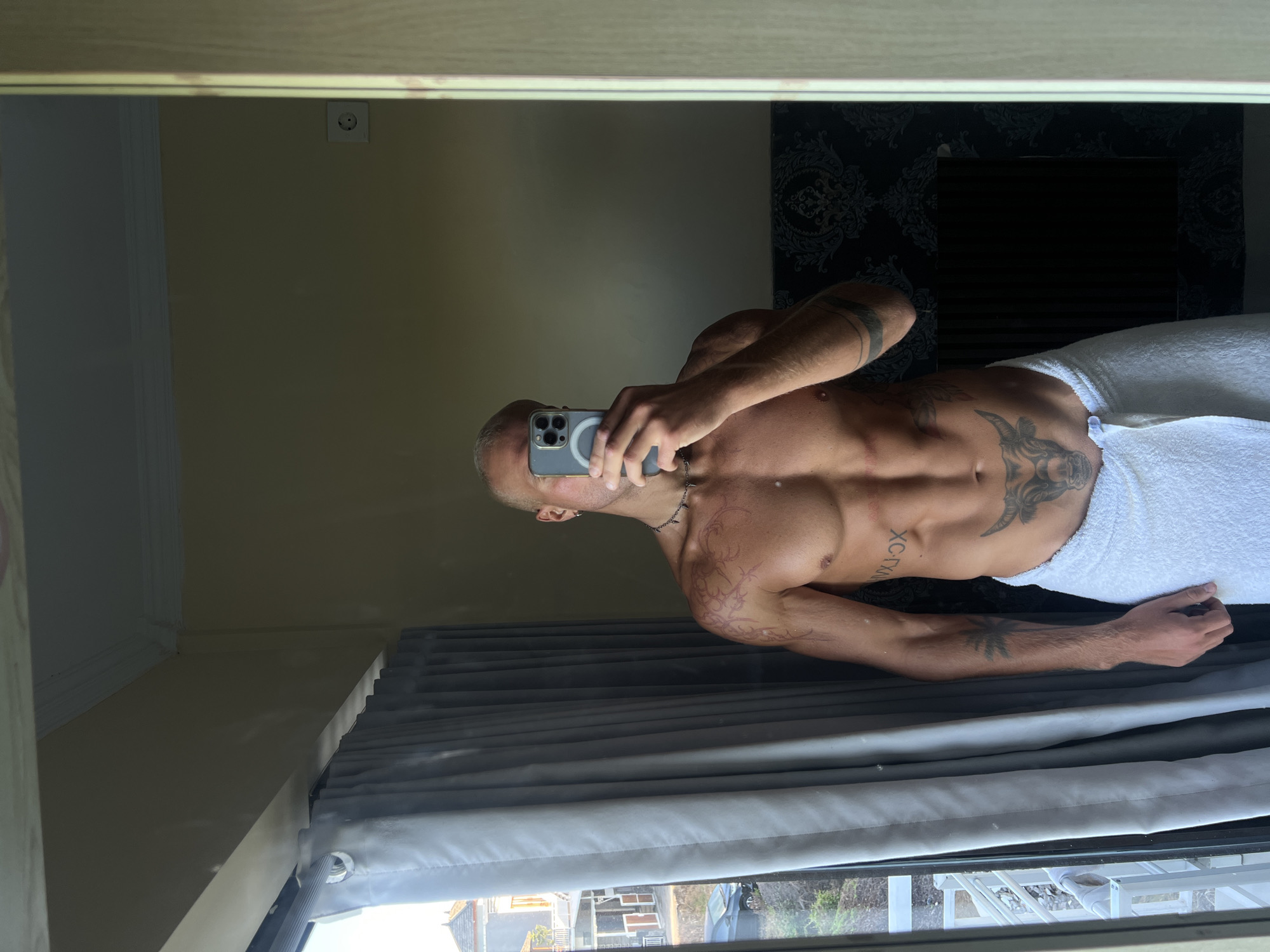 A man is taking a mirror selfie in what appears to be a bathroom in North Kuta, Indonesia. He is shirtless and wearing a white towel around his waist. He has several tattoos on his body, including a bull on his abdomen and tribal designs on his arm and chest. He is holding a phone up to take the picture. Through a window to his left, a street scene with houses and a car is visible. The room has yellow walls, a gray curtain, and a dark patterned wallpaper.