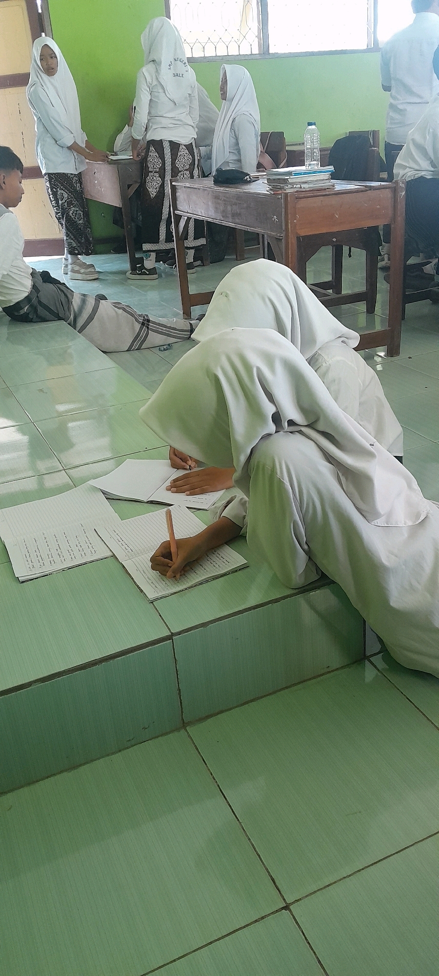 The image depicts an indoor scene, possibly a classroom, in Tahunan, Indonesia. A group of young individuals, predominantly female and wearing white hijabs and uniforms, are engaged in activities around wooden desks. One person is standing by a desk, seemingly interacting with another person. Two individuals are seated on the floor in the foreground, writing in notebooks. The floor is tiled in a green pattern, and the walls are painted green. Sunlight streams in from a window visible in the background. Objects such as notebooks, pens, a water bottle, and a backpack are present in the scene. The overall impression is one of an educational setting with students focused on their work.