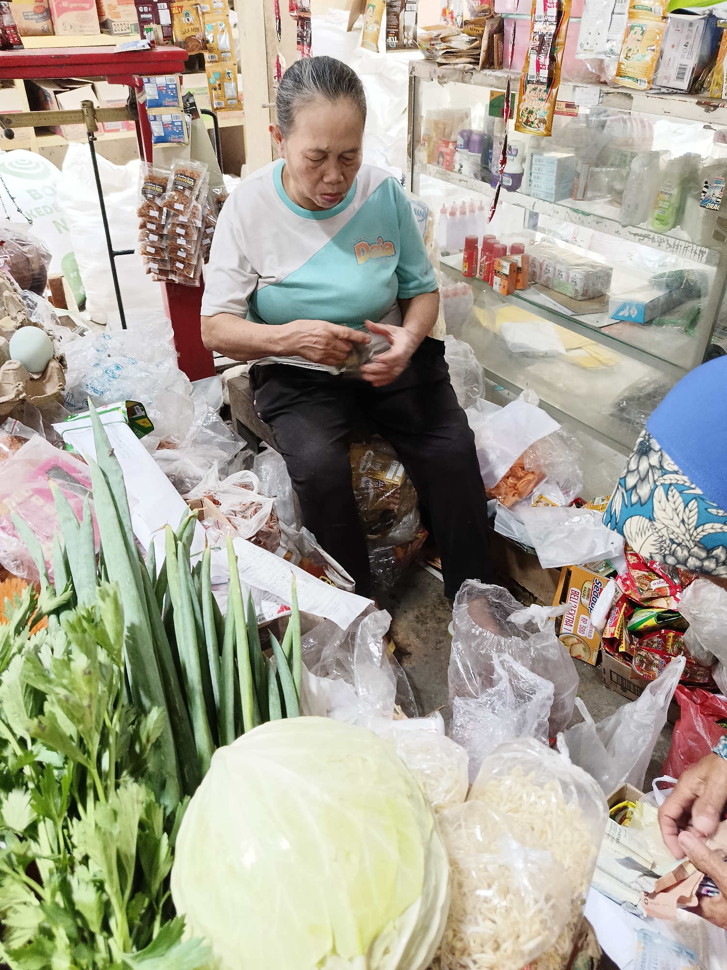 In Barukan, Indonesia, an elderly Indonesian woman with graying hair pulled back and closed eyes sits on a low stool amidst a cluttered market stall. She wears a white t-shirt with a teal color-blocked section across the chest and black pants. Her hands are engaged in handling what appears to be money and a small item.
The stall is densely packed with merchandise. To her left, a prominent display of fresh produce includes a large head of cabbage and a bunch of long green onions and celery. Various packaged goods, snacks, and other items are visible on shelves behind her and scattered around her on the floor, many contained in clear plastic bags. Some of these items include packets of dried snacks, instant noodles, and bottles of liquid. The overall impression is one of a busy, perhaps somewhat disorganized, local market environment.