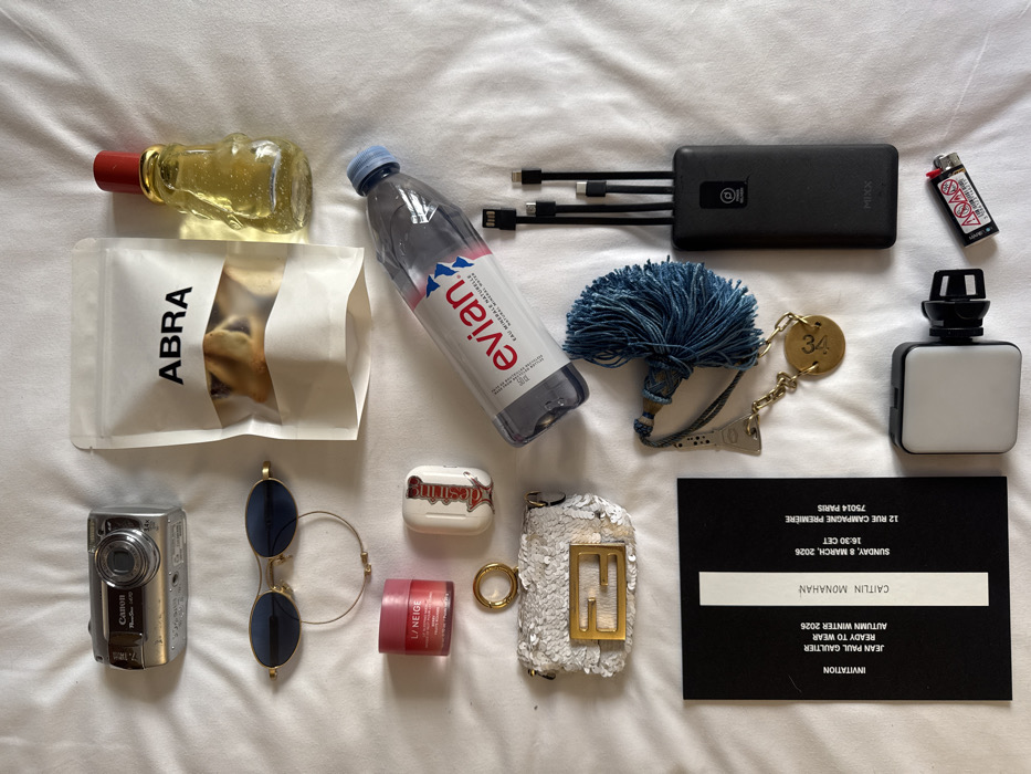 A flat lay composition displays various personal and event-related items arranged on a white, textured fabric surface. On the left, a silver Canon PowerShot SD1400 IS digital camera is positioned above gold-framed oval sunglasses with dark lenses, a thin gold-colored bracelet, a pink Laniege Lip Sleeping Mask container, and a white Apple AirPods case with "desiring" in red script. Below these is a small white sequined Fendi mini bag/keychain featuring a gold FF logo and a key ring. A black invitation card rests at the bottom left, detailing an event for JEAN PAUL GAULTIER READY TO WEAR AUTUMN WINTER 2026 for CAITLIN MONAHAN, scheduled for SUNDAY, 8 MARCH, 2026, at 16:30 CET, at 12 RUE CAMPAGNE PREMIÈRE, 75014 PARIS, France.
On the right side of the frame, a white pouch labeled "ABRA" contains baked goods, positioned above a small bottle with a red cap holding yellow glitter liquid, and a plastic Evian water bottle. Below these items are a dark blue tassel keychain attached to a gold chain with an Eiffel Tower charm and a round gold charm inscribed with "34." To its right is a black power bank with integrated USB-C, Lightning, and Micro-USB cables. At the bottom, a small white LED light on a black clip is next to a white disposable lighter.