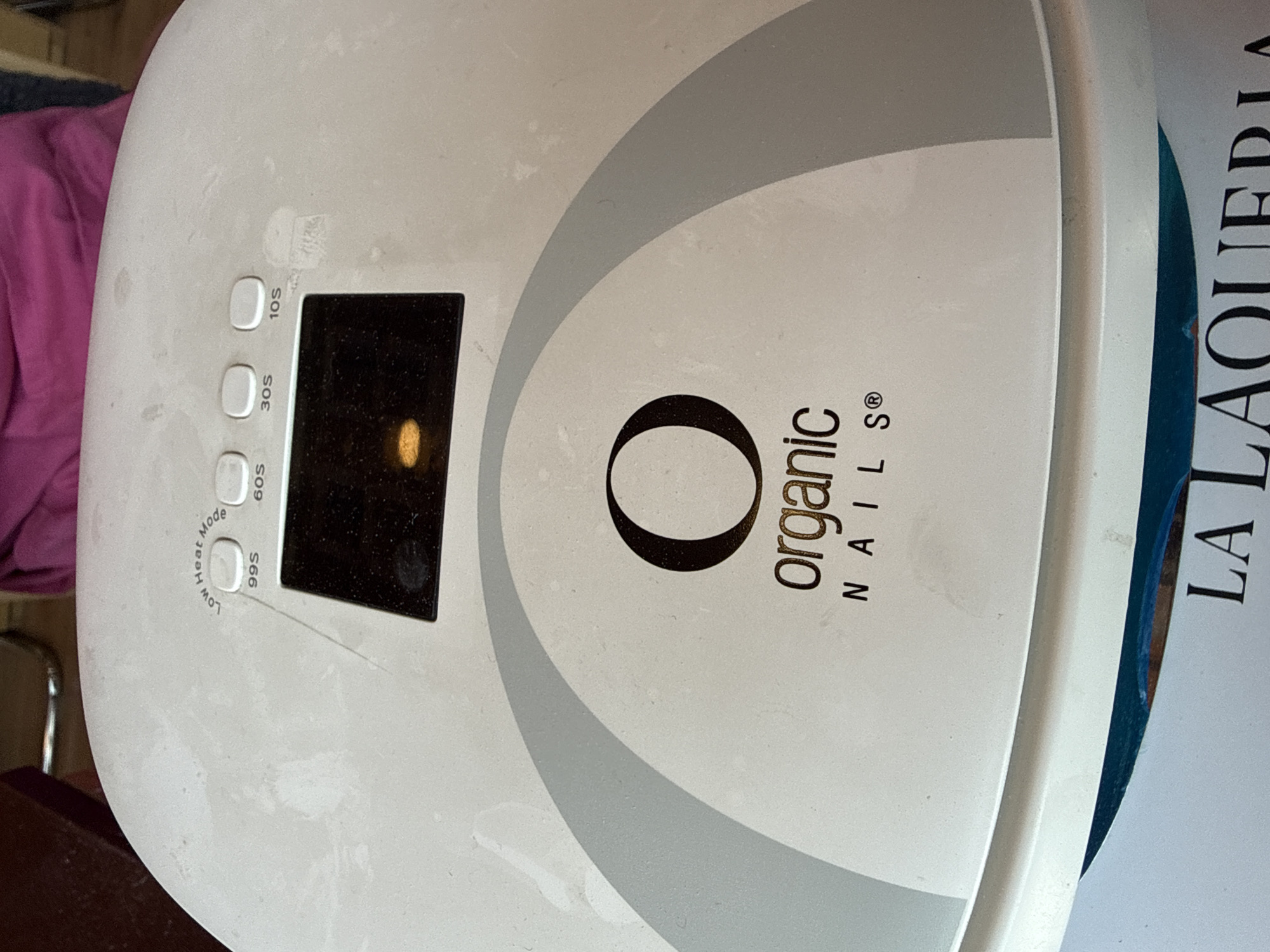 The image displays a white nail drying lamp from an overhead perspective, slightly rotated counter-clockwise. The lamp is the central and dominant subject, showing its top surface and control panel.
On the lamp's surface, the brand name "Organic NAILS®" is prominently displayed in black lettering. To the left of a rectangular digital display screen, there are four circular buttons. These buttons are labeled with different time settings: "10S", "30S", "60S", and "99S Low Heat Mode", indicating functions for varying drying times and a low heat option. The display itself shows a glowing orange or yellow light, likely indicating that the lamp is in operation or a light source within. The surface of the lamp appears slightly dusty or smudged, suggesting regular use.
In the background to the left, a piece of crumpled pink fabric is visible, possibly clothing or a towel. To the right, partially visible, is a white object with dark blue accents, on which the text "LA LAQUERIA" is written vertically in black, capital letters. This likely indicates a salon name or branding. The entire scene suggests an indoor setting, possibly a beauty salon or a personal nail care station in Puebla, Mexico, where "LA LAQUERIA" could be a local business. The lighting appears to be artificial. No people are visible.