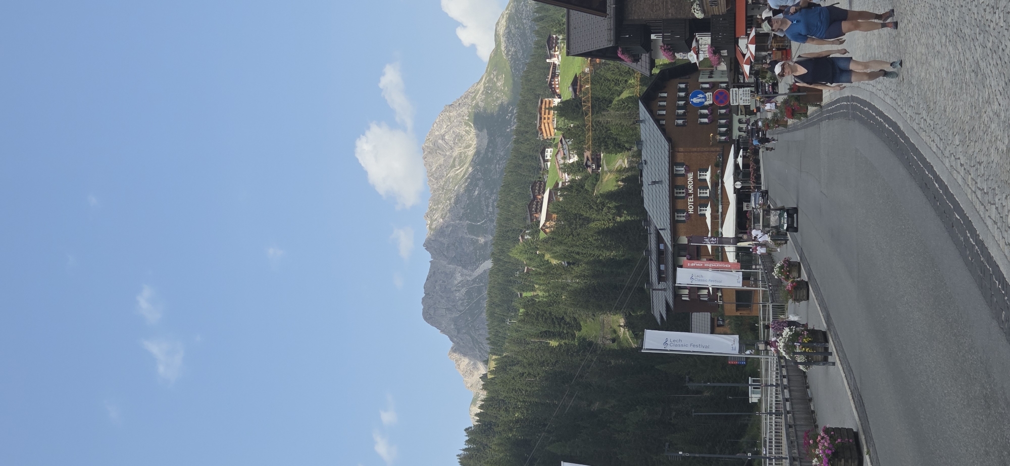 This is a vertical, eye-level shot of a street in Lech, Austria. In the foreground, two women are walking on a cobblestone sidewalk. The woman on the left is wearing a blue t-shirt, denim shorts, and a baseball cap, while the woman on the right is wearing a blue t-shirt, denim shorts, and a cap. The street is paved and curves to the right, with a few cars and a golf cart driving on it. Flower planters line the left side of the street, filled with colorful flowers.
Further down the street, several buildings are visible, including a hotel labeled "HOTEL KRONE." White umbrellas are set up outside the hotel. Flags with the text "Lech Classic Festival" are planted in front of the buildings. A ski lift can be seen in the background, ascending a tree-covered mountain. The mountain itself is rocky and rugged, with patches of snow. The sky is blue with scattered white clouds. The overall scene depicts a bustling summer day in a mountain town.