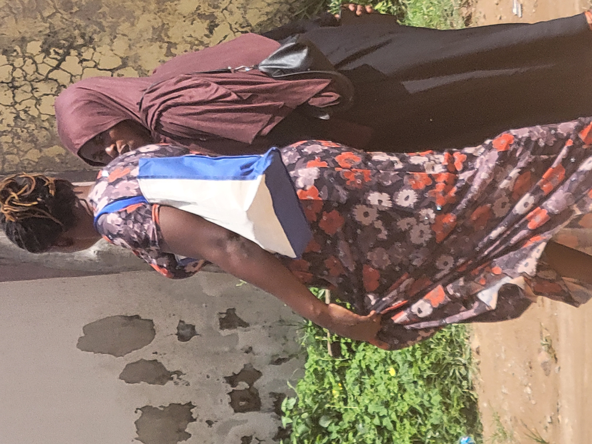 The image shows two women walking outdoors in Suleja, Nigeria. The woman in the foreground, on the left, is wearing a floral print dress with orange and white flowers on a dark background, and a white and blue bag over her shoulder. She is walking away from the camera, with her dress flowing around her legs. The second woman, on the right, is wearing a dark colored hijab and a long black dress. She is walking beside and slightly behind the first woman, with her head turned to her left, looking in the same direction as the first woman. She is carrying a black purse on her right shoulder. They are walking on a dirt path beside a wall that appears to be textured and peeling in places. Behind the wall, lush green foliage is visible. The lighting suggests it is daytime, likely sunny.