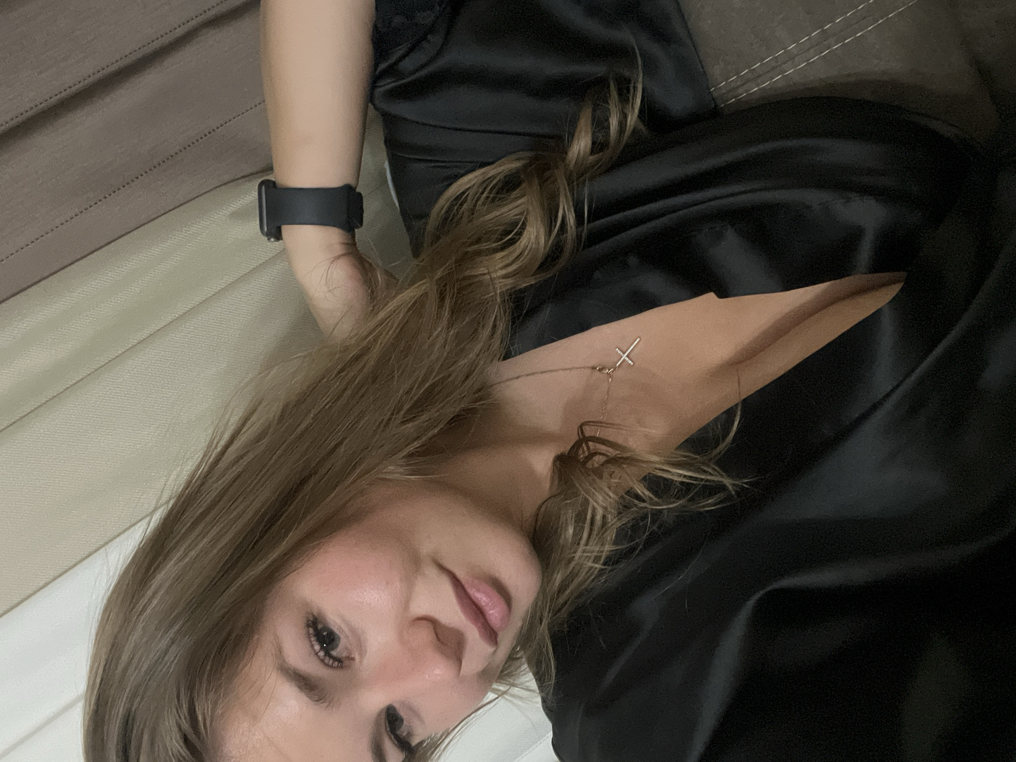 A young woman with shoulder-length blonde hair is lying on a brown and beige upholstered surface. She is wearing a black satin garment with a visible neckline. She has a black smart watch on her left wrist and a delicate necklace with a cross pendant. The location is not identifiable as Budva, Montenegro from the image provided.