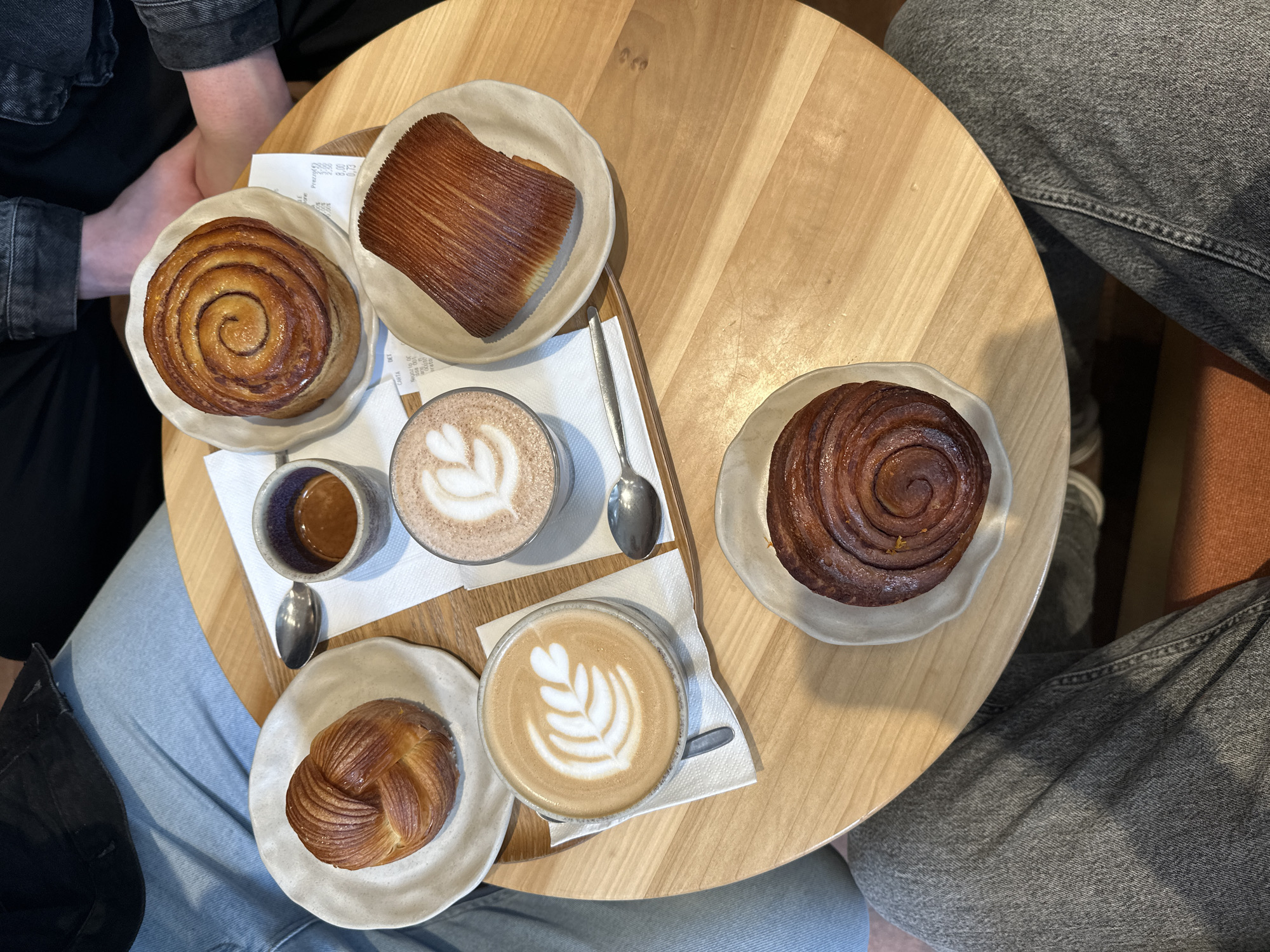 The image presents an overhead view of a light-colored wooden round table in a cafe, laden with coffee and pastries, with two individuals partially visible around it.
On the table are four distinct pastries: a large, golden-brown spiral pastry resembling a cinnamon bun on a light ceramic plate; a rectangular pastry with prominent, crisp layers, possibly a kouign-amann or similar laminated dough, on another light plate; a smaller, braided golden pastry on a third light plate; and a darker, chocolate-colored spiral pastry on a fourth matching plate.
Beverages include two lattes served in clear glasses, both featuring delicate white latte art of leafy designs. A small, dark ceramic cup contains an espresso shot. Several silver spoons and white paper napkins are placed among the items.
Partial views of two people are visible at the table. One person's arms, dressed in a dark denim jacket and black top, are seen near the cinnamon bun. The other person's legs, clad in light blue jeans and dark grey jeans, are visible around the table's edge. The scene suggests a relaxed cafe visit, likely during the daytime, given the bright appearance.
A crumpled paper receipt is visible on the table. Legible text includes "PREZZO (R)", itemized prices such as "3,50", "2,50", "8,00", and "0,75", along with "CARTA", "DET.", "N. 000101", and a timestamp "ORA 10:16", indicating a morning transaction. The overall atmosphere is casual, portraying a typical coffee and pastry experience in a cafe setting, likely in Milan, Italy.