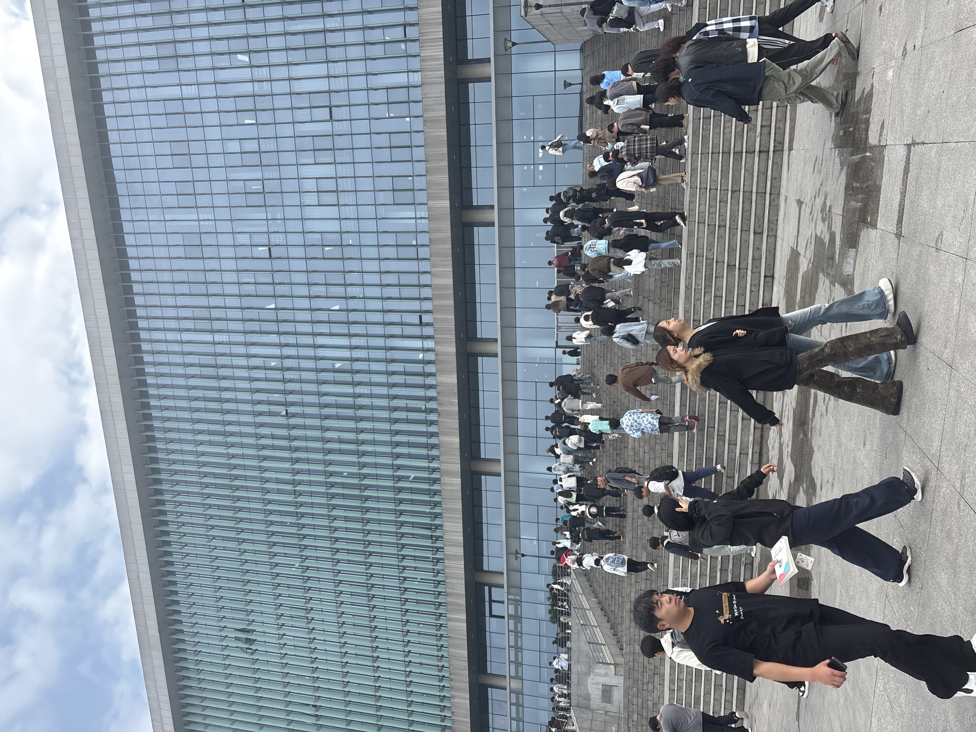 The image captures a bustling outdoor scene in Jiangjin, China, in front of a modern building with a predominantly glass facade. A large group of people, seemingly students, are present on a wide set of steps leading up to the building. Most individuals are casually dressed in a variety of clothing styles. They are walking or standing, some appearing to be engaged in conversation. The architecture is contemporary, with the building's reflective surface mirroring the sky and clouds. The setting suggests an educational institution or a public space near one.