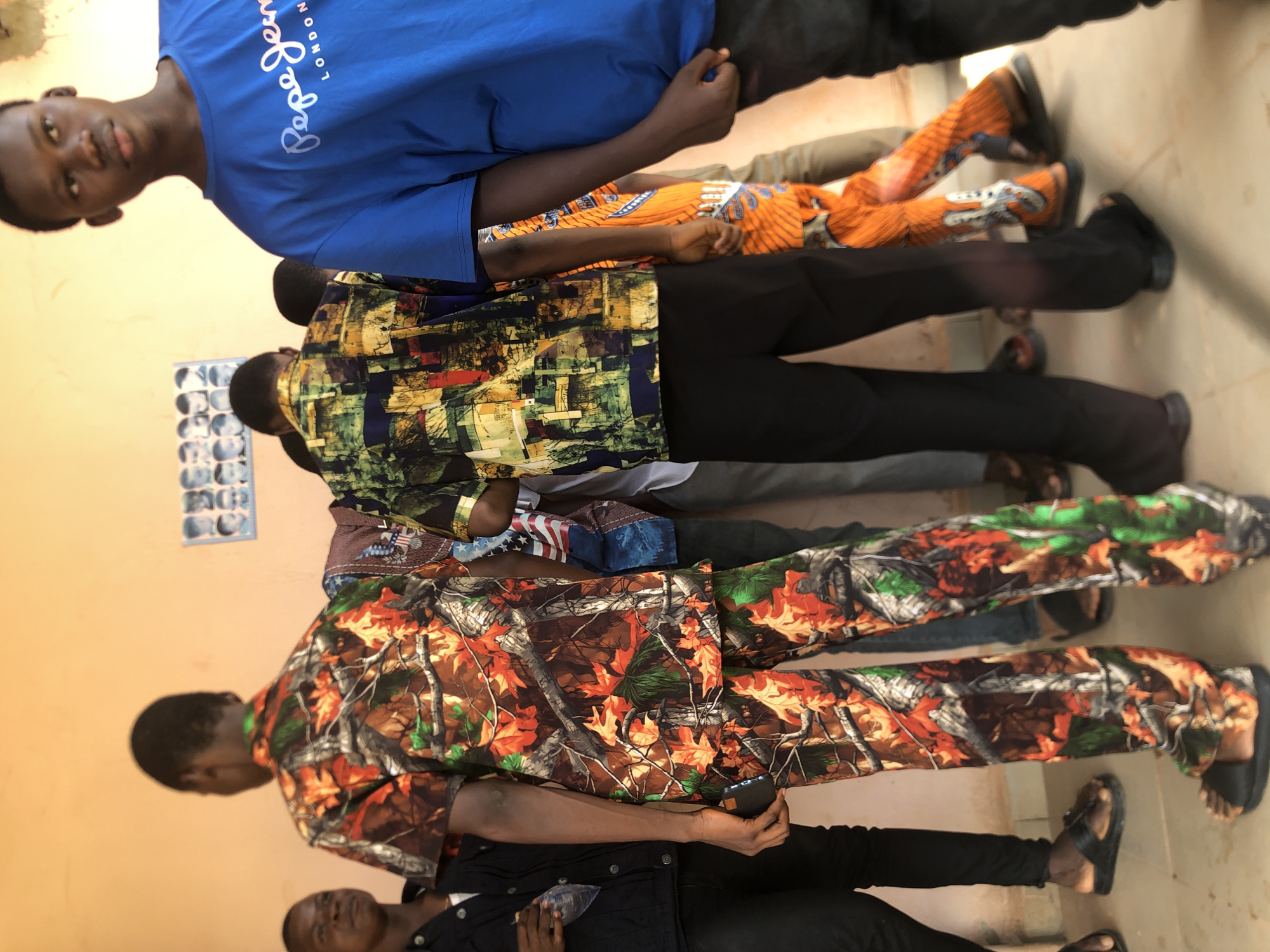 The image shows a group of men standing indoors against an orange wall in Nkwelle Ezunaka, Nigeria. From top to bottom: A man in a blue t-shirt is partially visible. The next man is wearing a colourful orange and blue patterned shirt. The next man is wearing a busy multicoloured shirt with black trousers. The next man is wearing a shirt composed of the American flag. The next man is wearing a shirt patterned with autumnal leaves and colours. The bottom man is wearing a dark shirt and trousers. Each man wears sandals or shoes. A printed document is taped on the wall behind the first men.