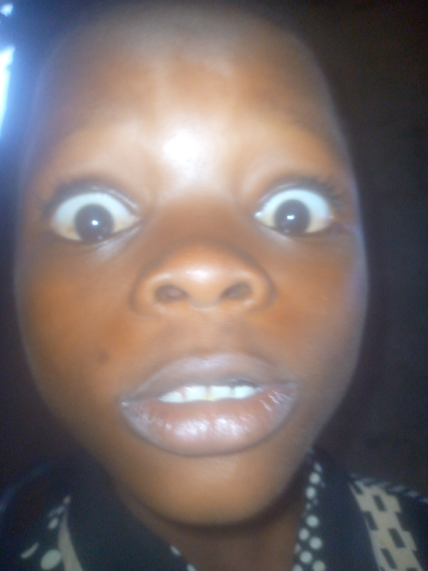 A close-up, head-on shot captures the face of a young child with dark skin. The child is looking directly at the camera with a wide-eyed, perhaps surprised or playful, expression. Their eyes are open unusually wide, revealing a significant amount of white around the dark irises, with a clear reflection of a light source visible in both pupils. The child's mouth is slightly agape, showing several white teeth.
The lighting in the image is somewhat dim overall, but the face is illuminated, possibly by a camera flash, which creates a bright highlight on the forehead and eyes. The background is dark and out of focus, offering no discernible details about the setting, though the overall appearance suggests an indoor environment or low-light conditions.
A portion of a patterned garment, possibly a shirt or top, is visible across the bottom of the frame. It features light-colored polka dots or squares against a dark fabric. There is no visible text or branding in the image. The time of day is not ascertainable from the image. No specific location cues are present. The child's emotion appears to be one of mild surprise or a deliberate comical pose.