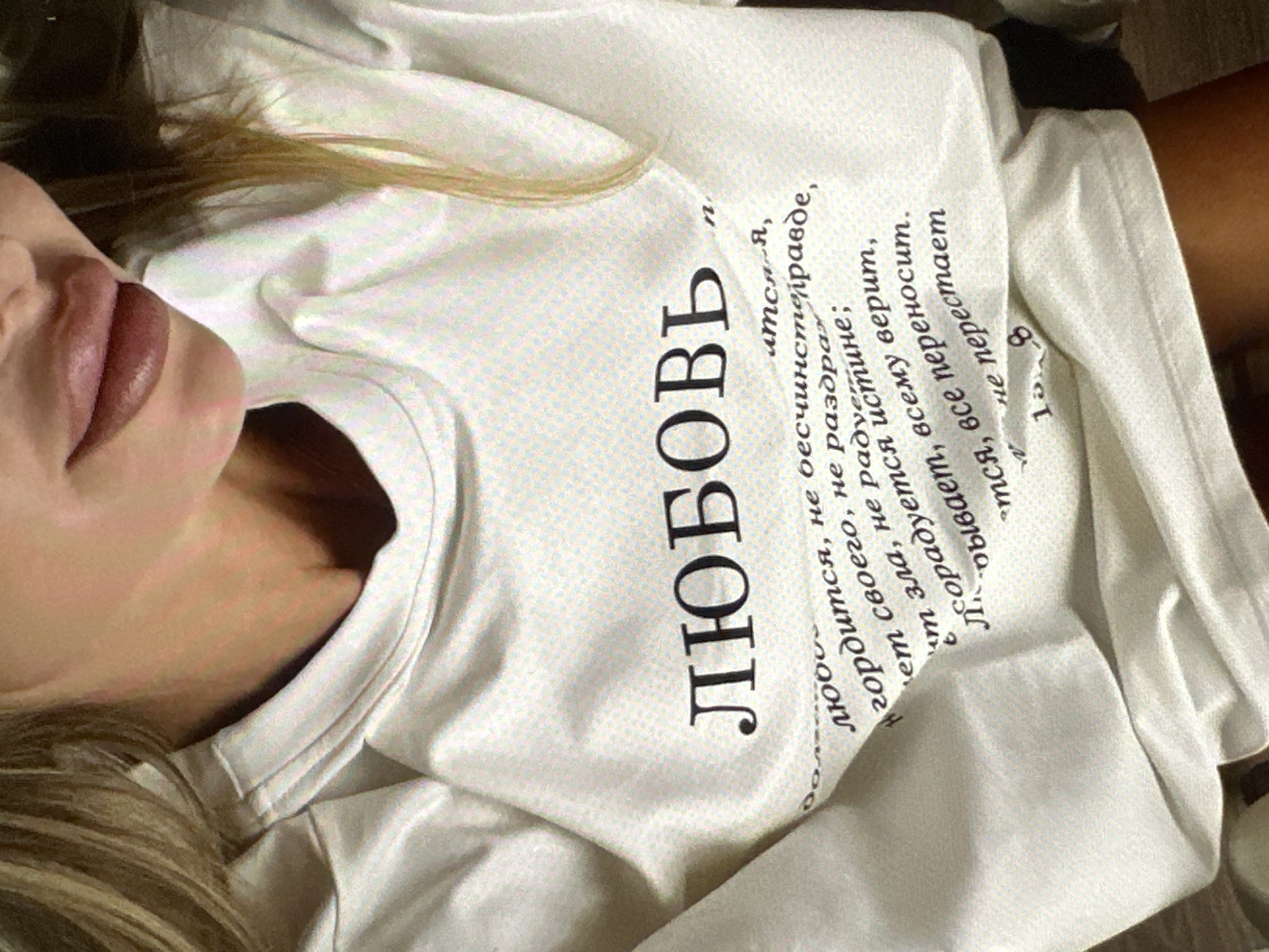A woman is lying down, presumably indoors, wearing a white t-shirt with Cyrillic text printed on it. The largest word on the shirt, oriented vertically, is "ЛЮБОВЬ" (Lyubov), which translates to "Love." The rest of the text appears to be a longer passage or verse in Cyrillic script. Her hair is blond, and her lips are visible. The setting is undeterminable from the given image. There's no information in the image to suggest that it was taken in Yerevan, Armenia.