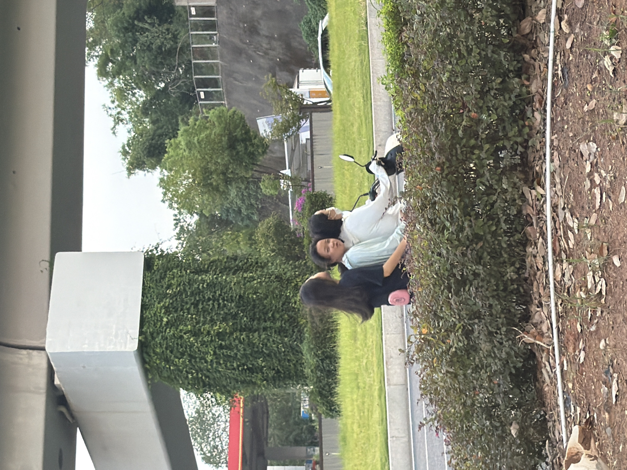 In Jiangjin, China, three young women are riding on a scooter. The woman in the back is wearing a black shirt and has long dark hair. The woman in the middle is wearing a light blue and white hooded jacket and has black hair. The woman in the front is wearing a white jacket and a mask. They are on a road with a grassy median. Behind the median is a large concrete structure covered in ivy, and further in the background are trees and a hill with a fence. To the left, a gas station canopy is visible. The foreground consists of a low hedge and dry leaves on the ground.
