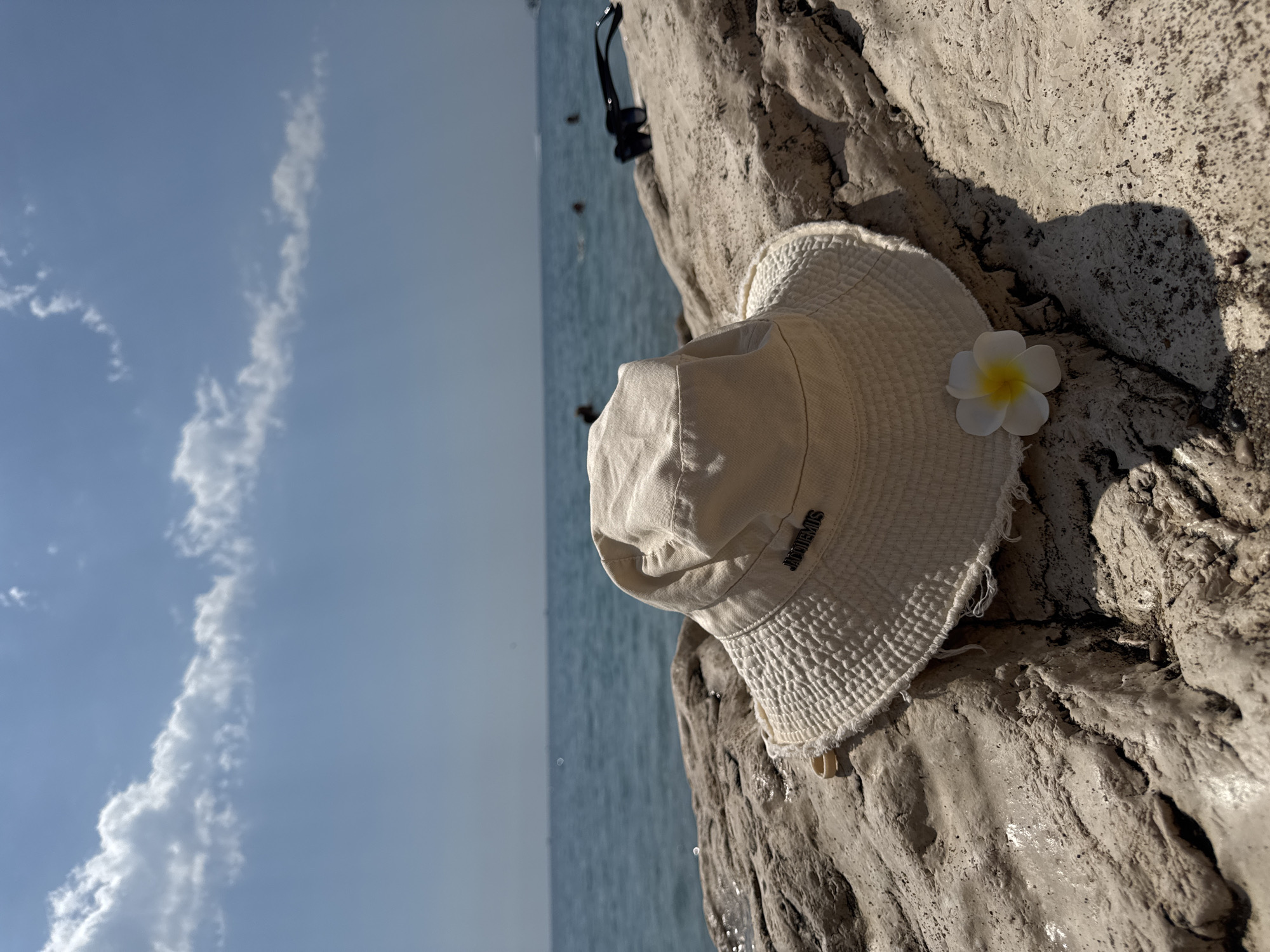 A close-up shot focuses on a cream-colored bucket hat placed on a textured, light-colored rock formation. The hat has a frayed edge and a small metal logo on the front that reads "JACQUEMUS." To the right of the hat, a white plumeria flower with a yellow center rests on the rock. In the background, the blue sea stretches to the horizon under a bright blue sky with scattered white clouds. Several people are visible swimming or wading in the water in the distance. A pair of dark sunglasses are also placed on the rocks, further back and to the right of the hat. The scene appears to be set on a rocky shoreline, likely in the city of Kemer, Turkey, given the clear water and sunny weather typical of the Mediterranean coast.